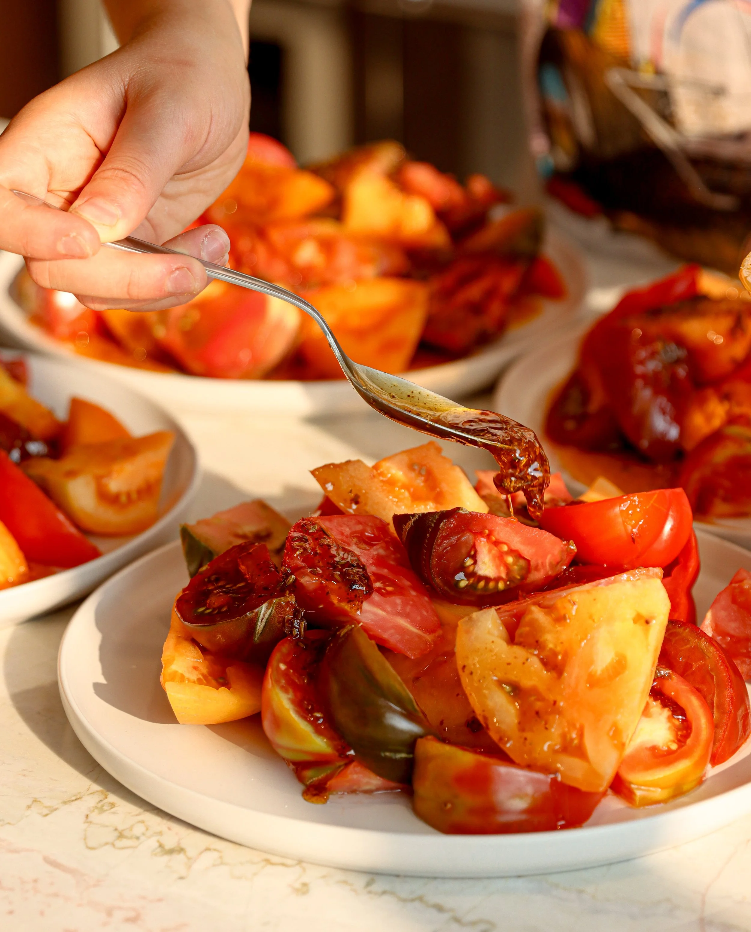 A close-up of a hand holding a spoon with barbecue sauce, with a platter of chopped fruits, including tomatoes and peaches, in the background.