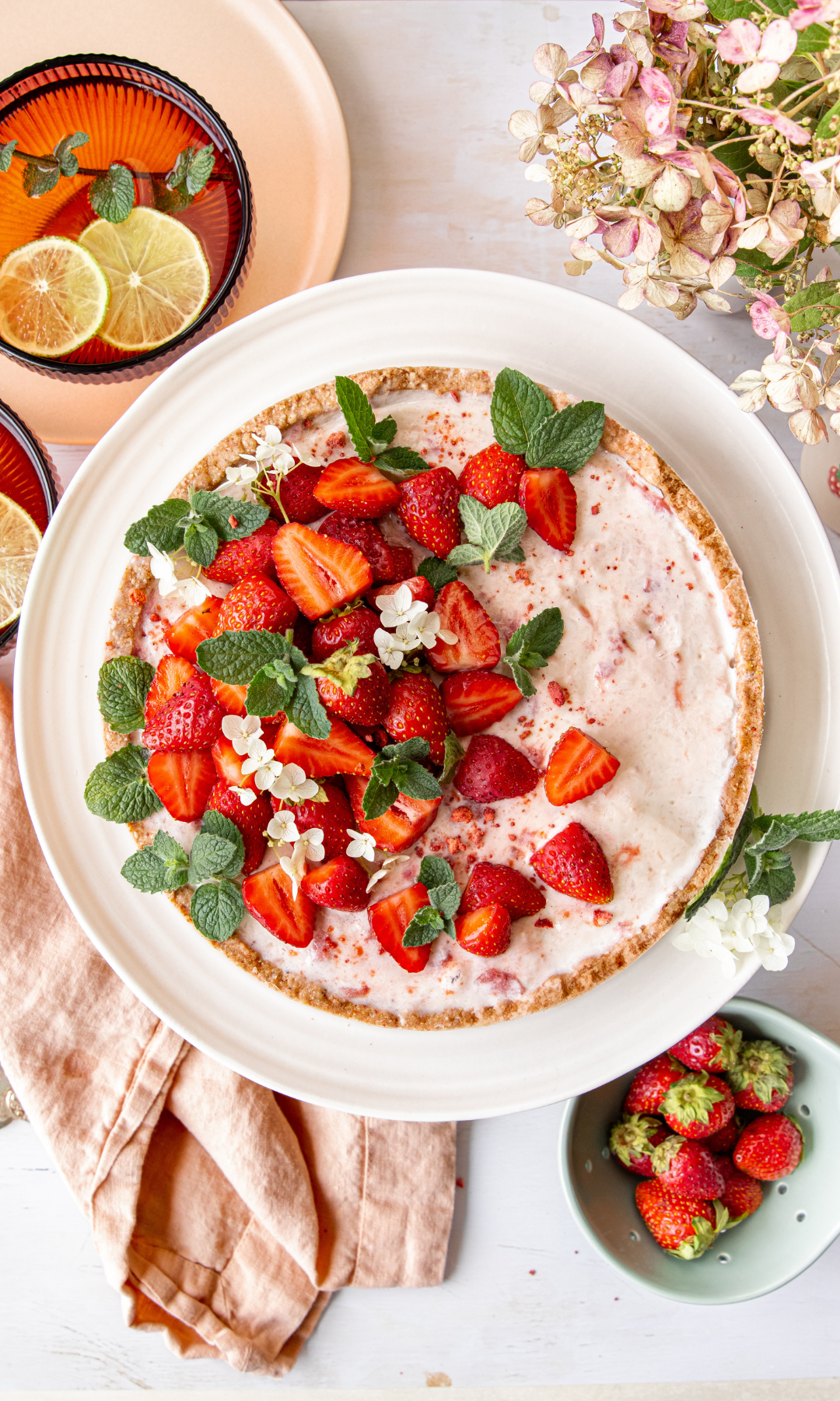 A strawberry tart topped with fresh strawberries, mint leaves, and small white flowers, served on a white plate, with side bowls of strawberries and glasses of lemon-infused water, and a pink cloth napkin.