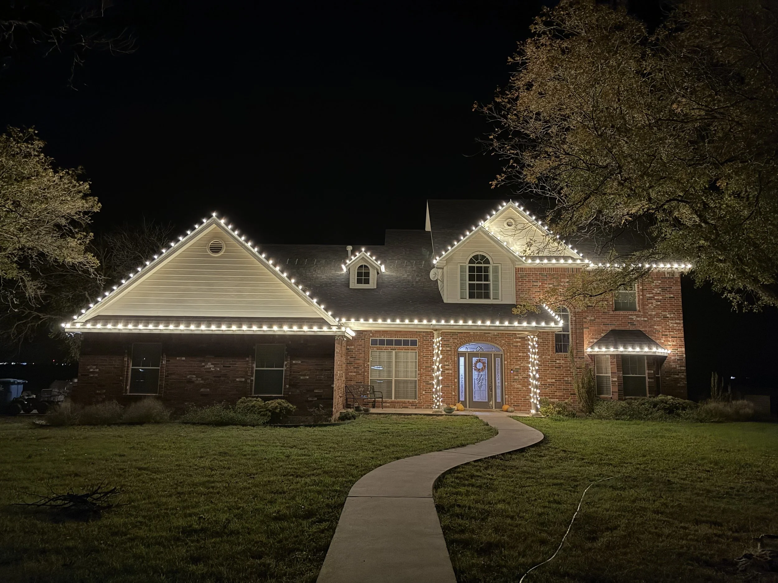 A two-story brick house illuminated with string lights along the roof edges at night. The house has a front door decorated with a wreath, topped with arched windows on the second floor, and is surrounded by trees with some leaves fallen. A winding concrete walkway leads to the front door, and pumpkins are placed near the entrance.
