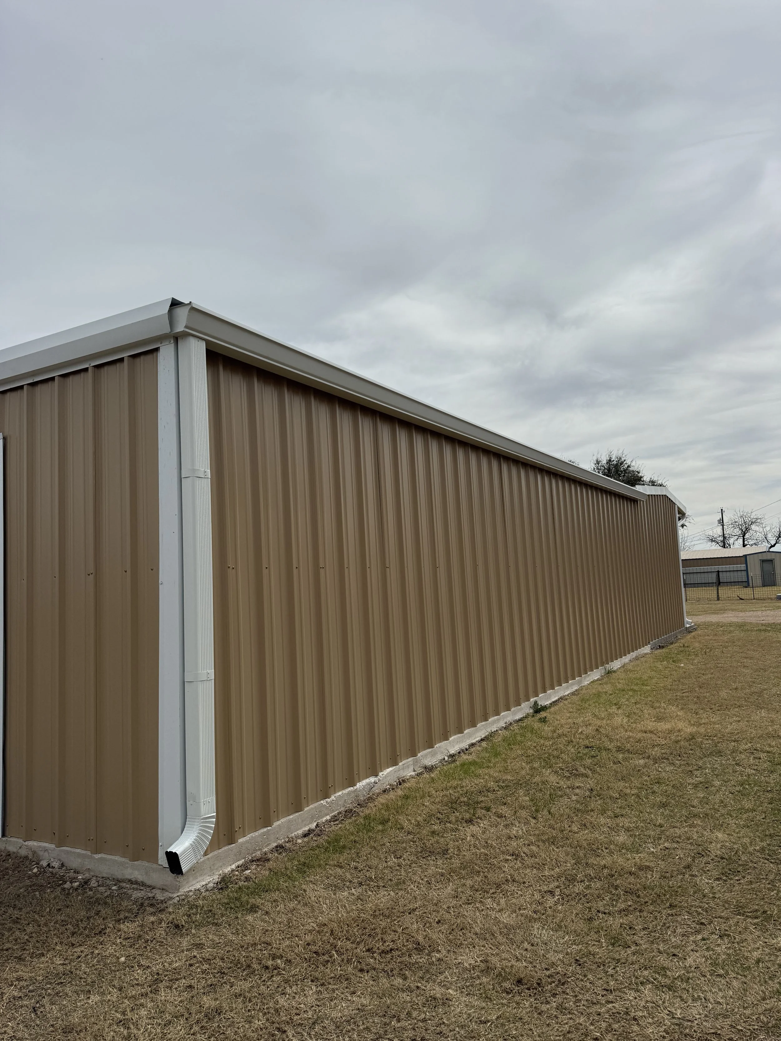 The side of a beige metal building with a white gutter and downspout, on a patchy grass yard under an overcast sky.
