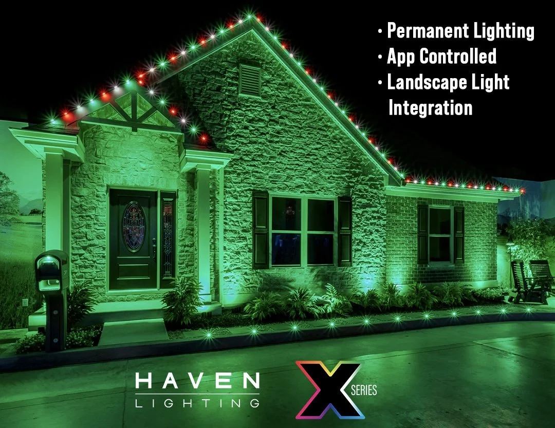A house decorated with green and red holiday lights along the roofline. The house features brick and stone siding with lit windows and a front door. Surrounding plants are illuminated in green. On the right, outdoor furniture is visible. The image promotes Haven Lighting's X Series landscape lighting, emphasizing permanent, app-controlled landscape light integration.