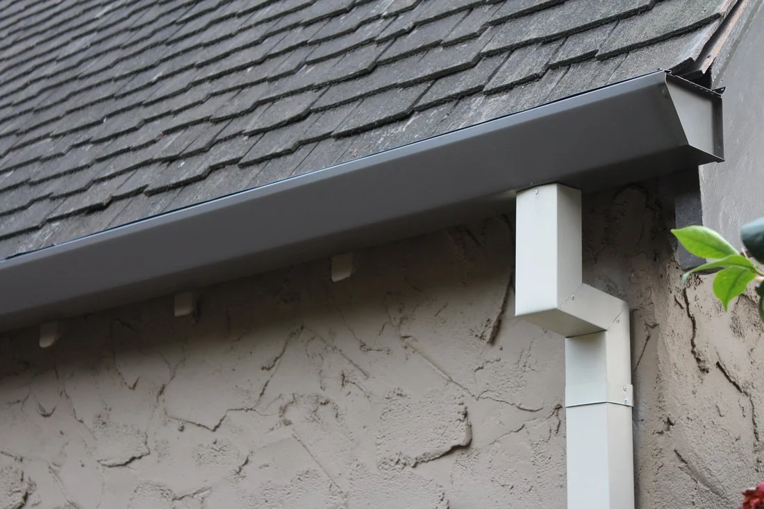Close-up of a house's exterior with gray metal flashing, a gray roof shingle, and a white gutter downspout attached to a beige stucco wall.