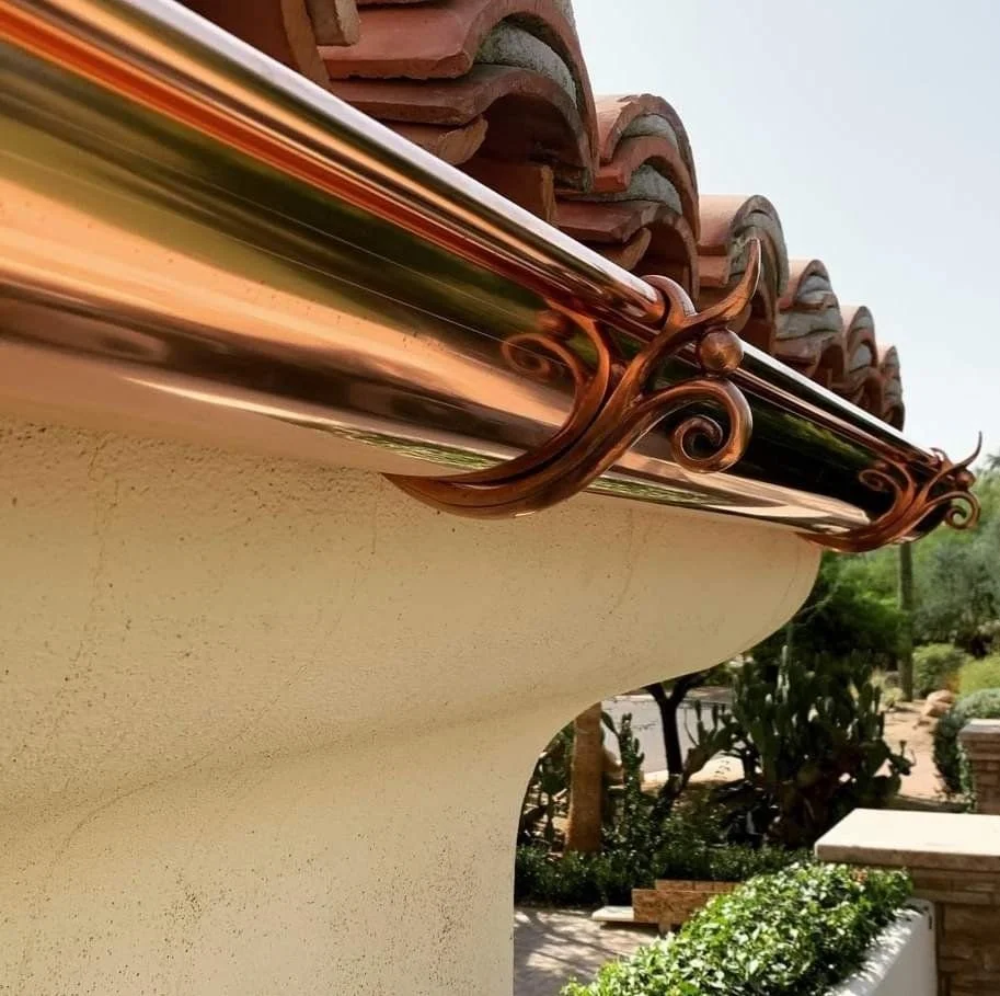 Close-up of a house's eaves with decorative copper brackets and copper gutter, with a beige stucco wall and a backyard with trees and shrubs in the background.