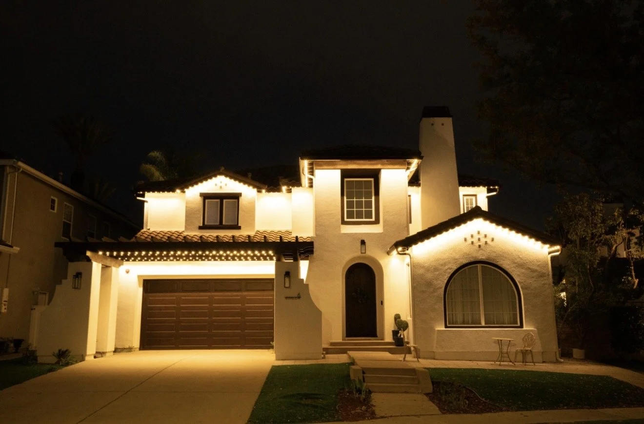 A two-story house illuminated with warm yellow holiday string lights at night, with a dark sky, trees, and greenery visible around it.