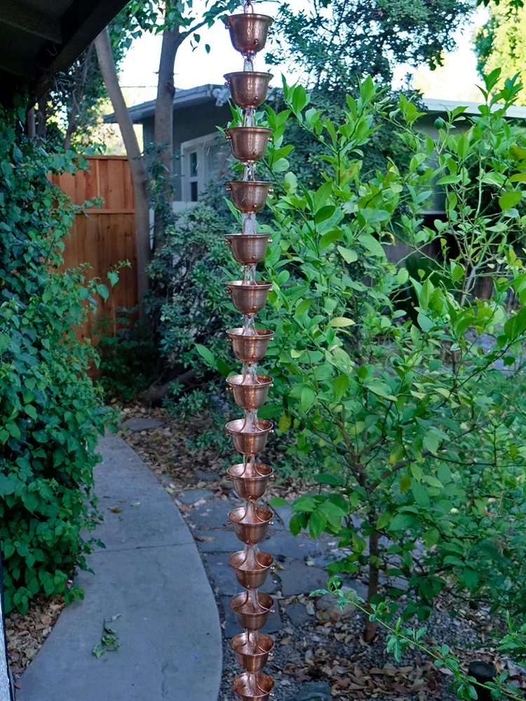 Copper water fountain sculpture with multiple small cups stacked vertically outdoors in a garden with bushes, trees, a concrete walkway, and a house in the background.