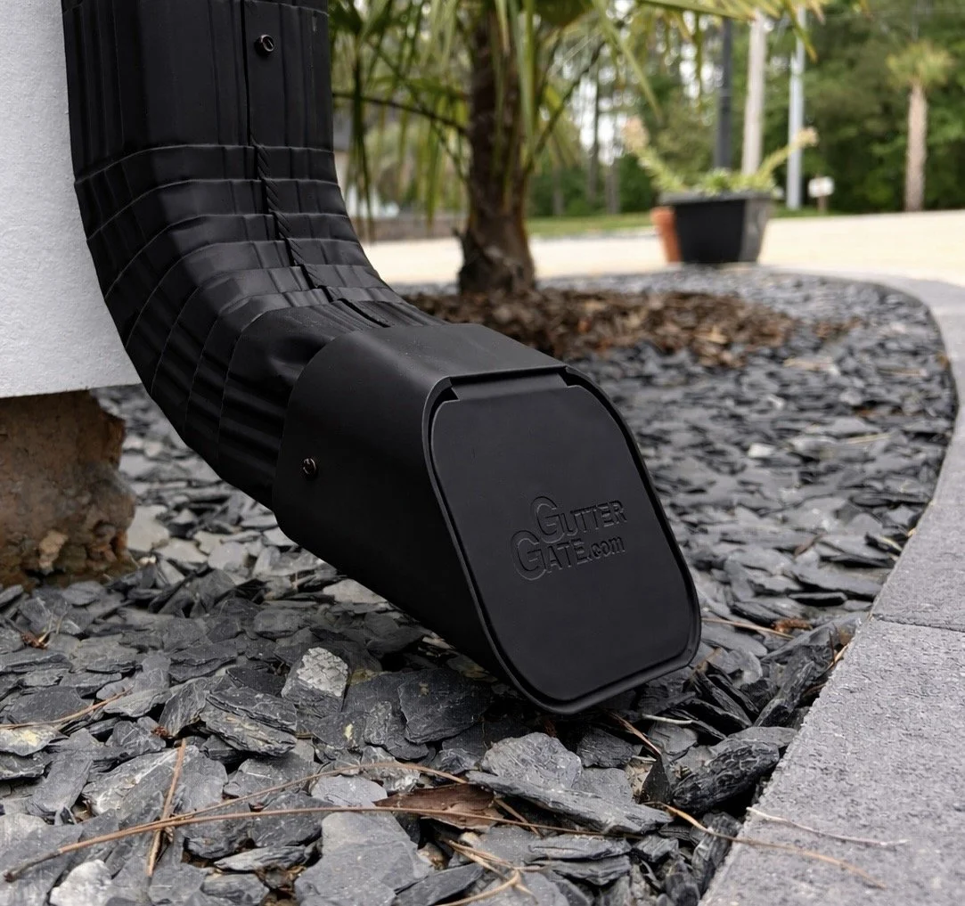 Black gutter drain extension attached to downspout, labeled GutterGATE.com, on a building, with slate gravel and a landscaped yard in the background.