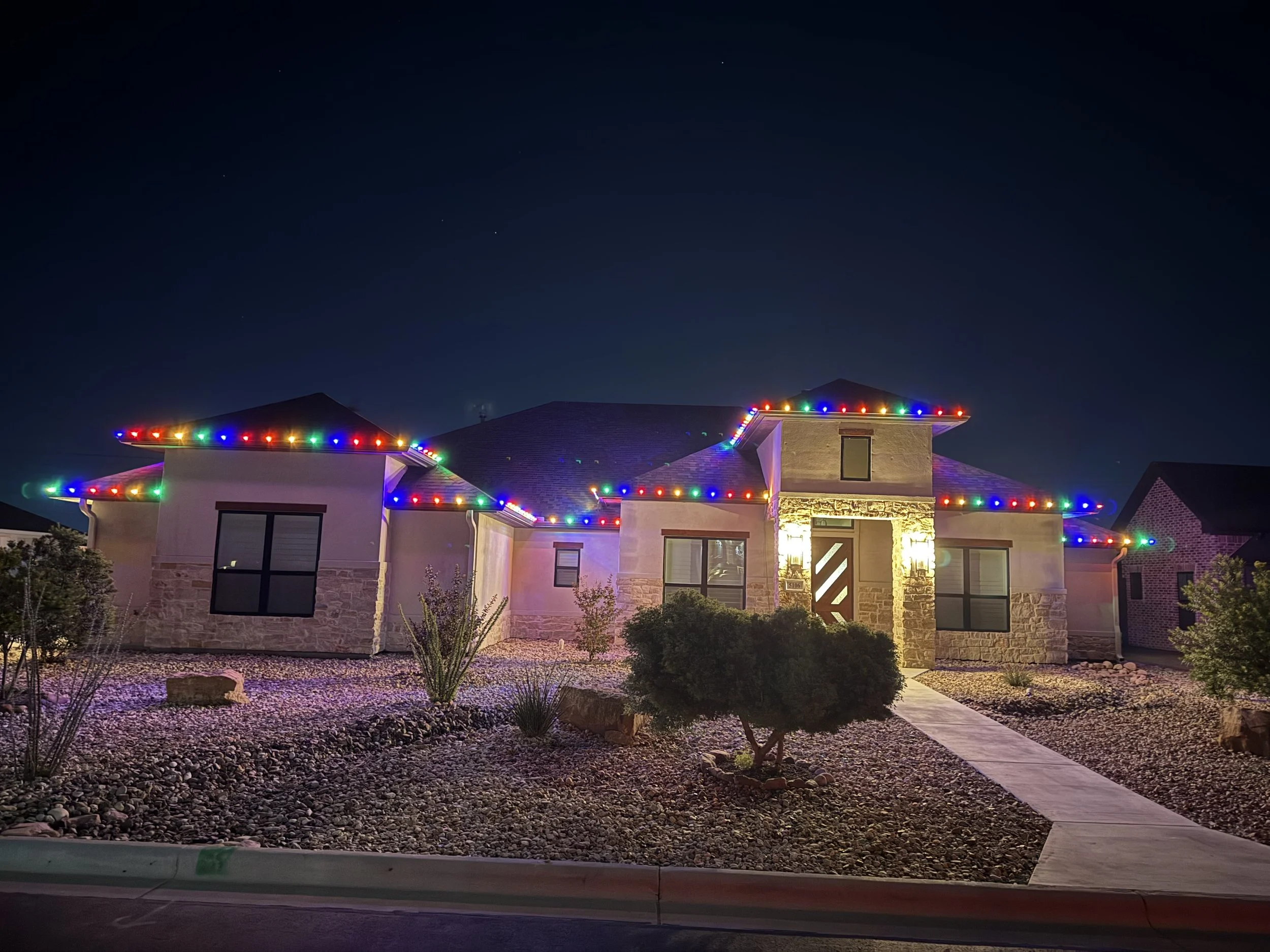 A modern house decorated with colorful Christmas lights outlining the roof. The house is at night with a dark sky and visible stars. The front yard has rocks, bushes, and a sidewalk leading to the front door.