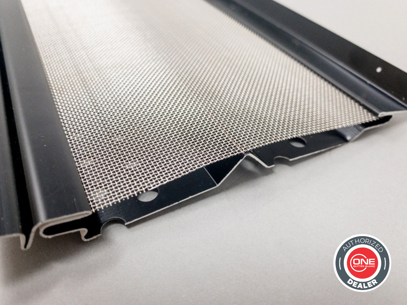 Close-up of a metal gutter guard with a fine mesh screen, mounted on a black frame. A logo in the bottom right corner reads 'Authorized Dealer' and 'One Gutter Guard'.
