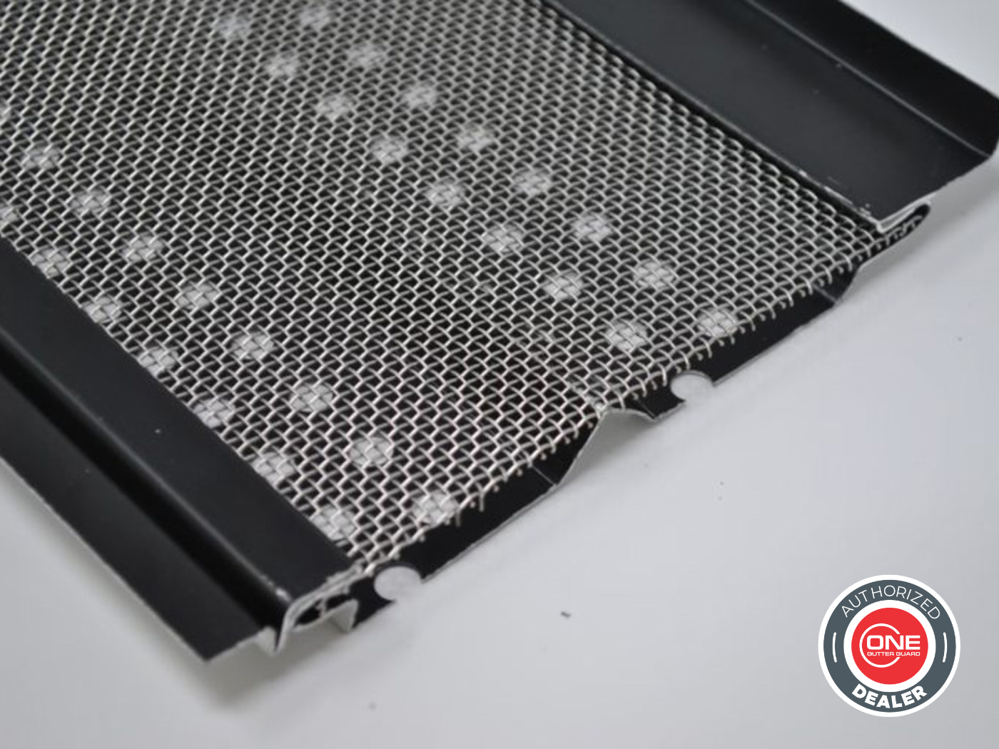 Close-up of a black metal gutter guard with a mesh design, placed on a white surface with a partially visible black frame on the right side.