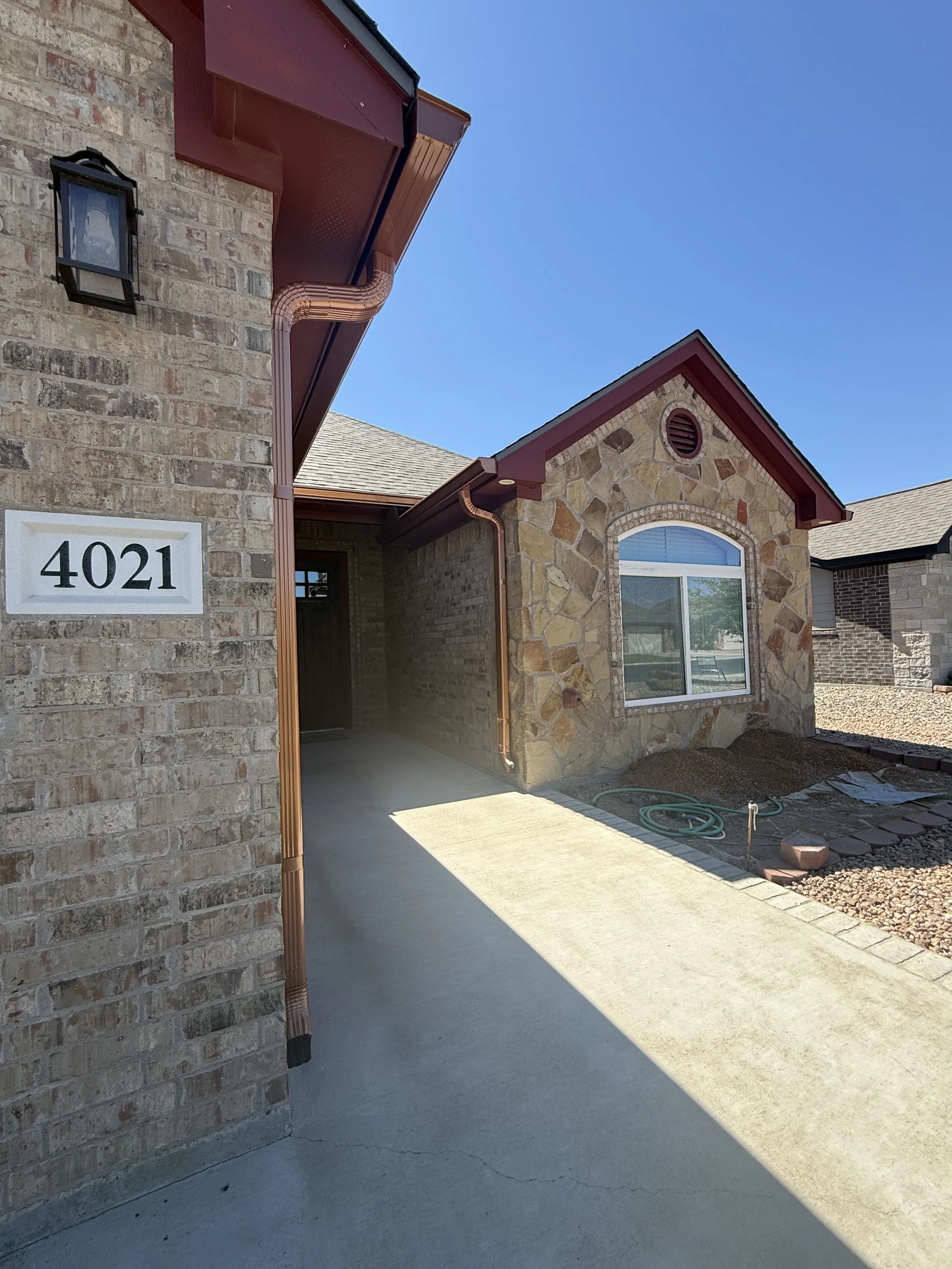 Front porch of a house with brick and stone exterior, a large window, and house number 4021. There is a concrete driveway and some construction materials on the ground. The sky is clear and blue.