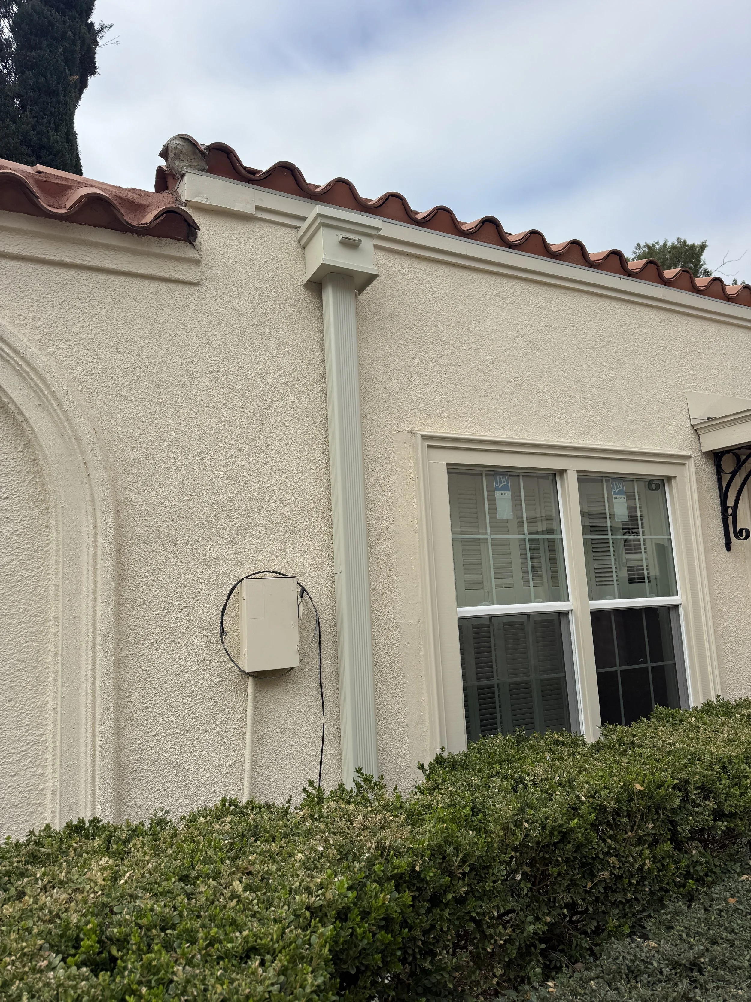 Exterior of a house with a stucco wall, window, trash can, and shrubbery in front, with a red tile roof and a gutter system.