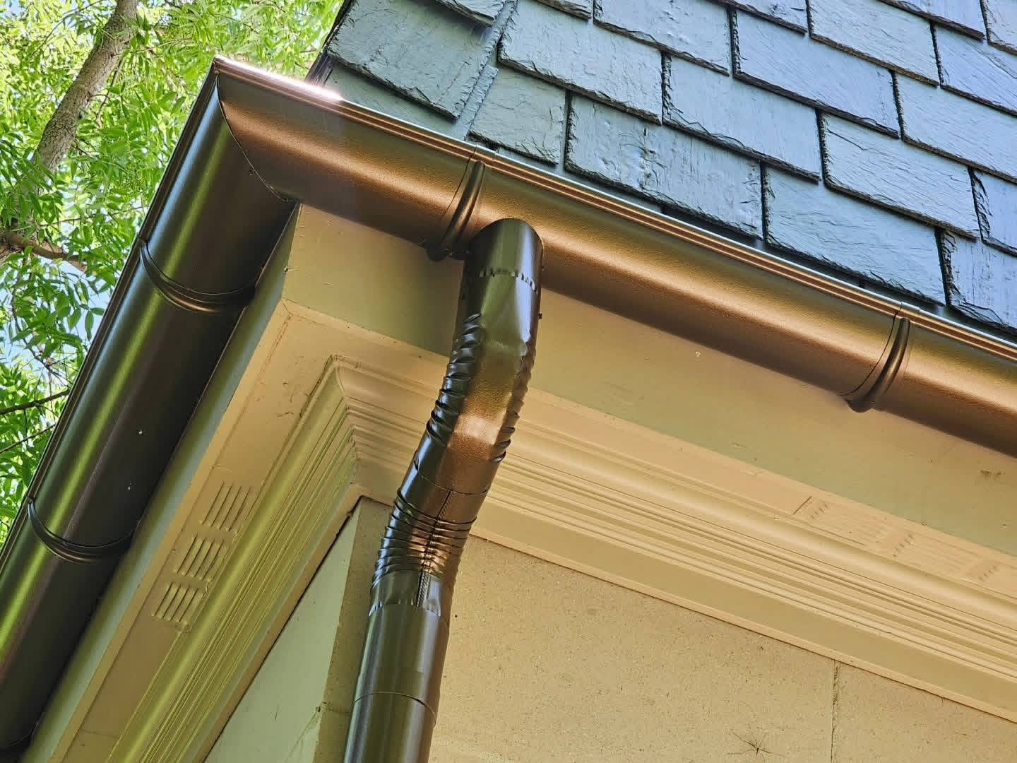 Close-up view of a house gutter system with a downspout attached, situated beneath a house overhang, with greenery and tree branches in the background.