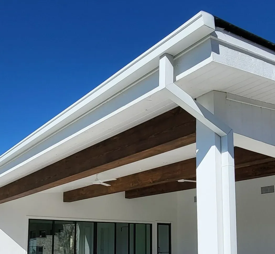 Close-up of the corner of a modern house exterior with white walls, large wooden beams on the ceiling, and black-framed glass doors, against a bright blue sky.