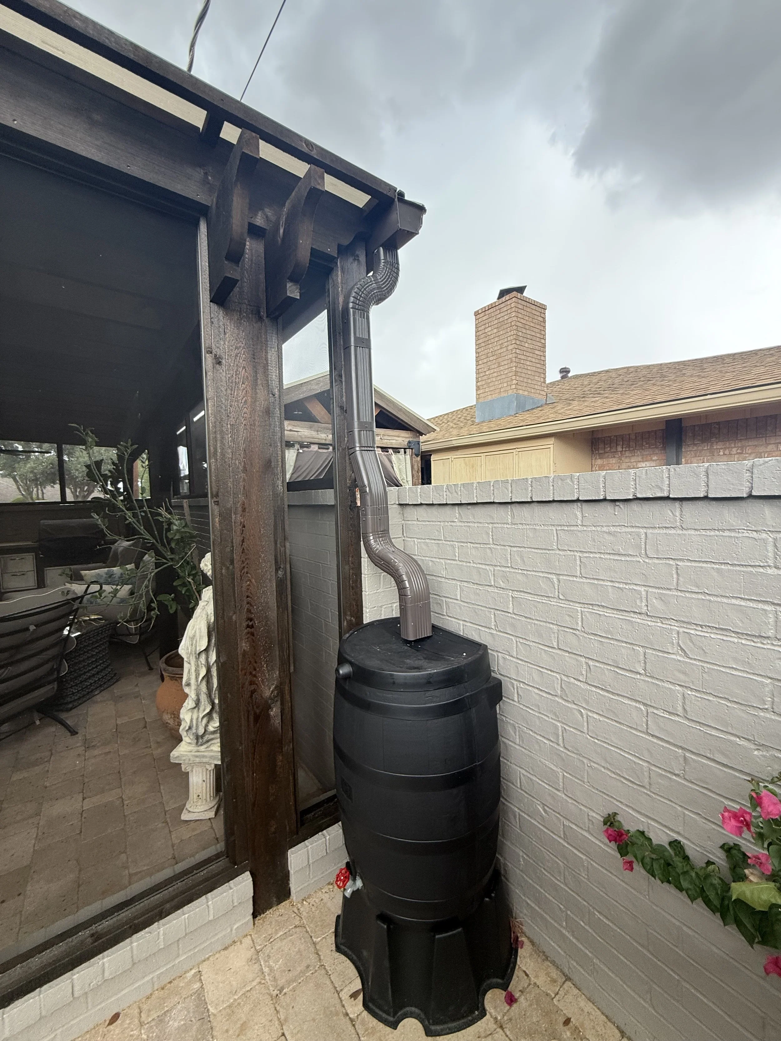 A backyard patio scene showing a black rain barrel with a black lid, a wooden beam supporting the patio roof, a rain chain, a brick chimney on a house with a beige exterior wall and a shingle roof, and pink flowers growing in a pot.
