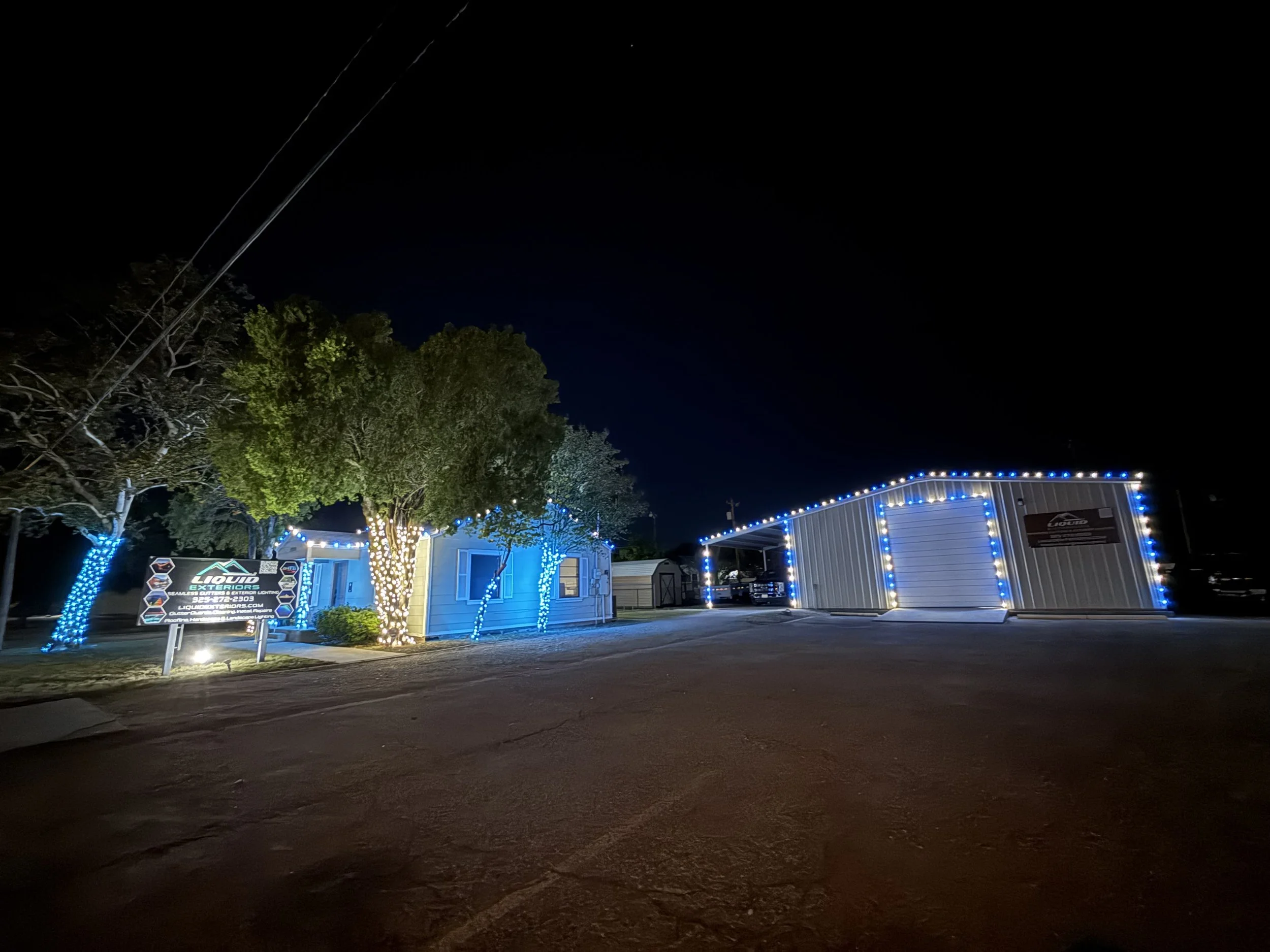 Night view of a building decorated with blue string lights, green trees with lights, a sign for Liquid Exteriors, and a clear sky with a visible star.