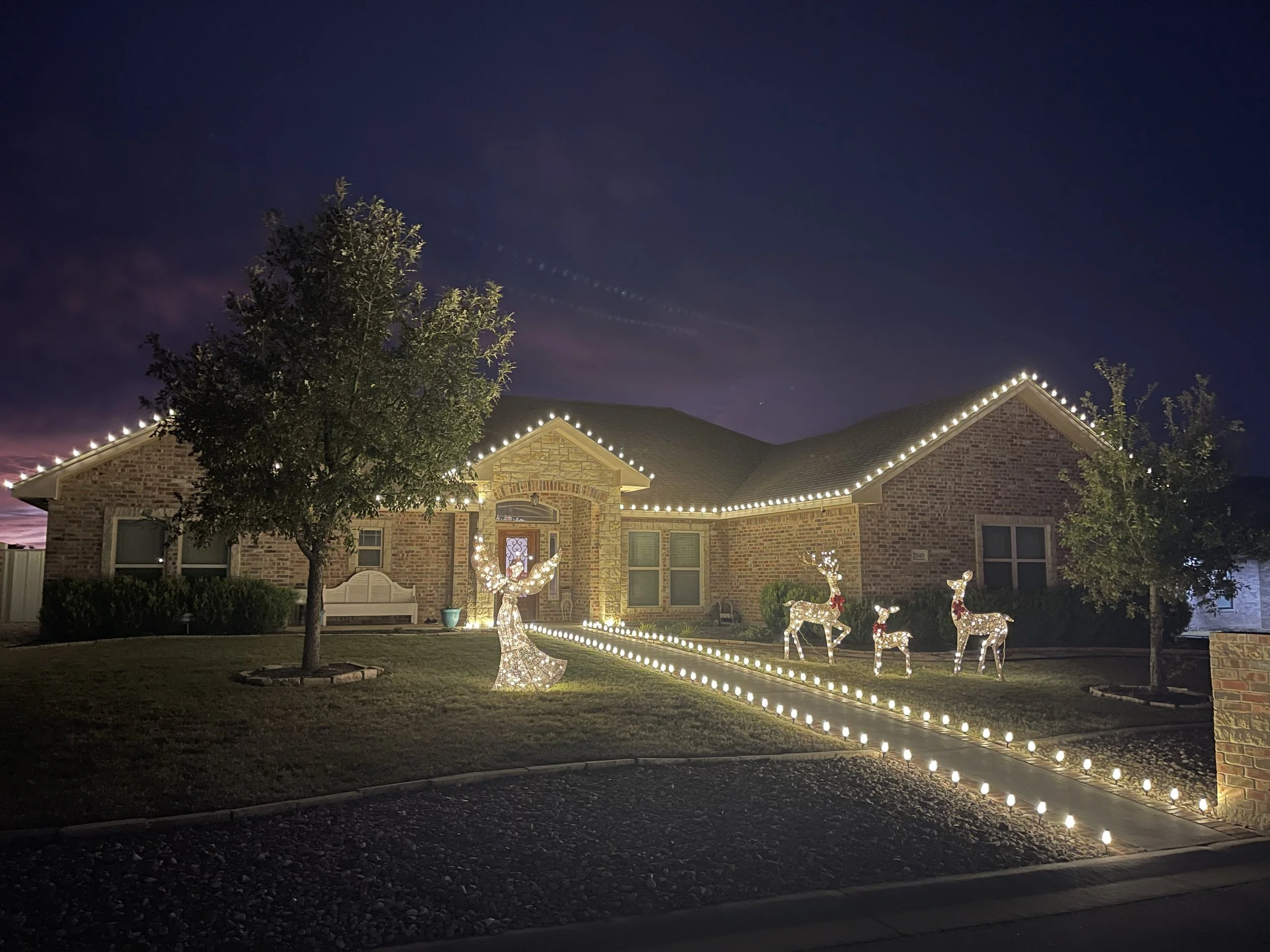 A house decorated with Christmas lights. The house has a brick exterior, a gabled roof, and a front porch with a window above the door. Christmas lights outline the roof and the front yard includes illuminated reindeer and a Christmas tree figure. There are trees and a sidewalk in the yard, and the photo is taken during the evening or night.