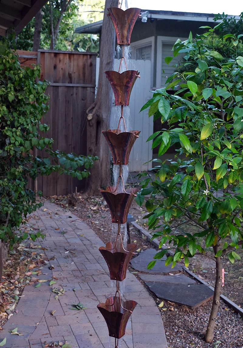 Outdoor garden scene with a vertical water fountain made of metal flower-shaped basins connected by copper tubing, surrounded by green bushes and a small tree.