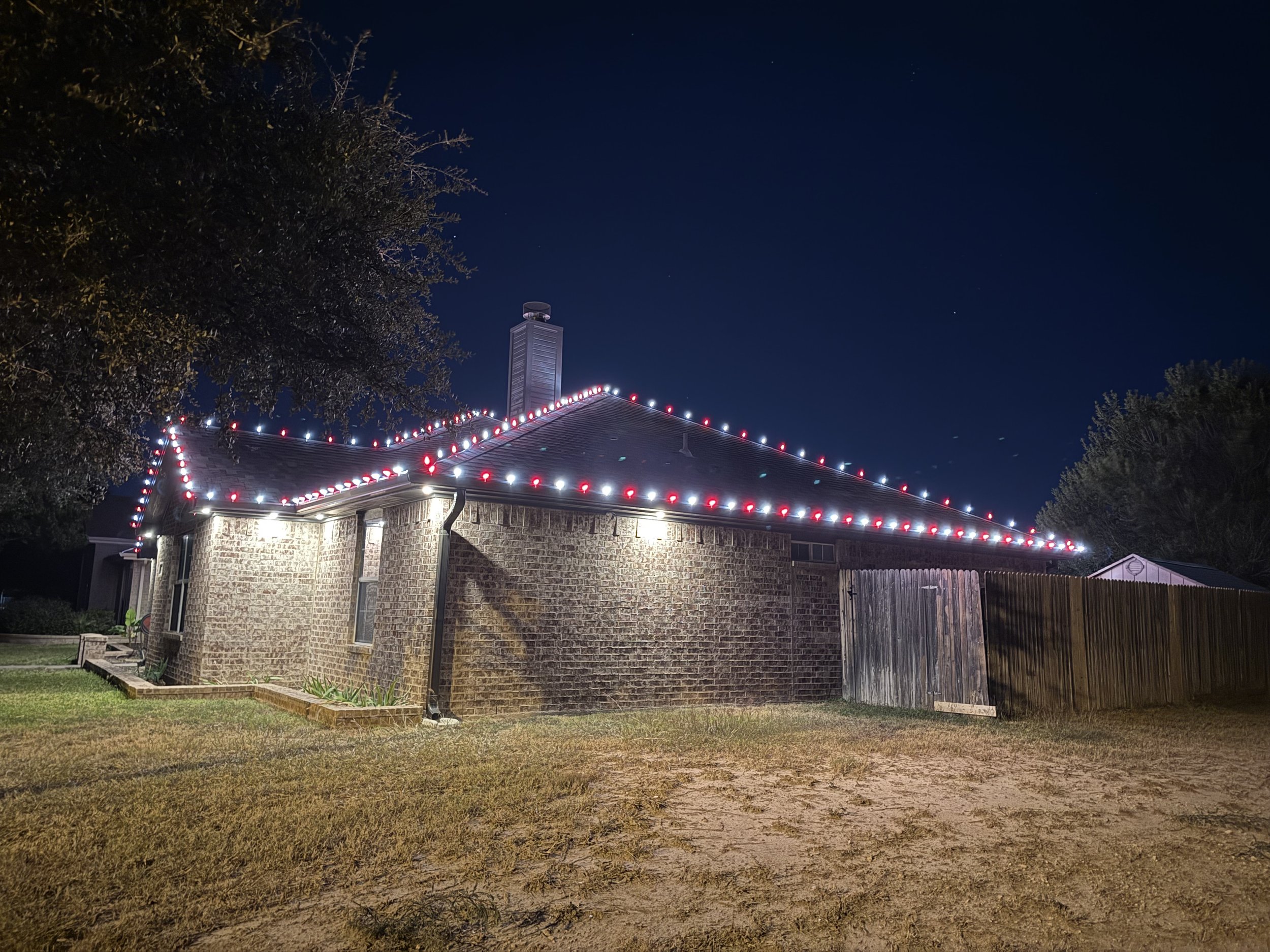 Nighttime view of a brick house decorated with red and white Christmas lights along the roofline, with a tree on the left and a wooden fence on the right, and a dark sky with some stars.