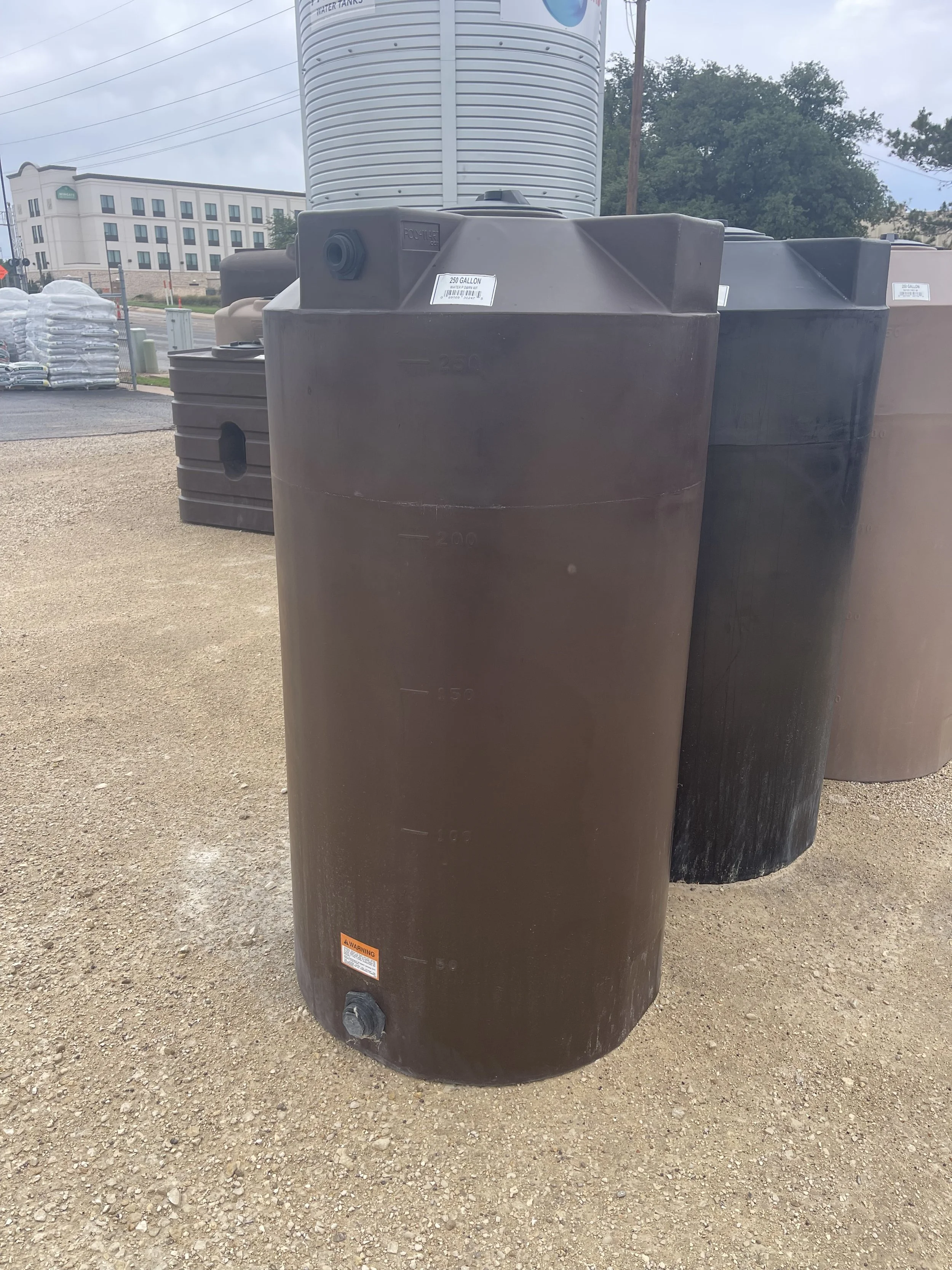 Large black water storage tank with a measurement slot and a warning label, placed on a gravel surface outdoors.