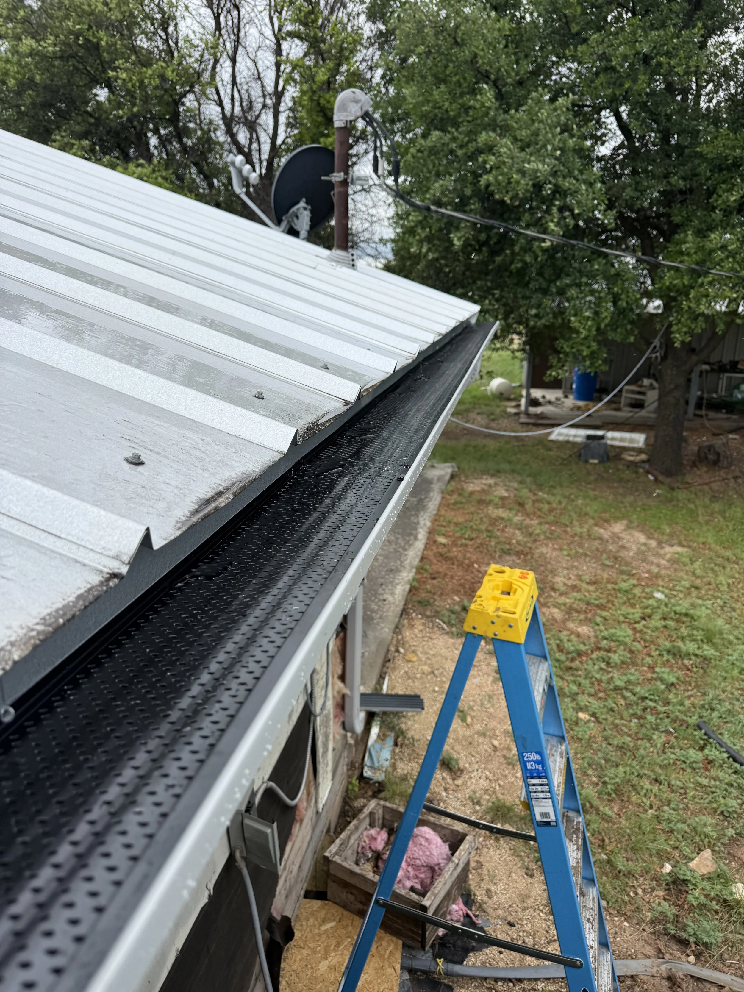Ladder and part of a building's roof with a metal surface, outdoor scene with trees, and construction or maintenance tools and equipment nearby.