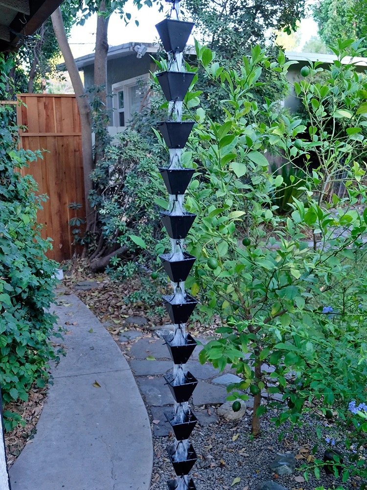 A vertical outdoor water fountain with black tiered bowls, placed in a garden with green foliage and a stone pathway.