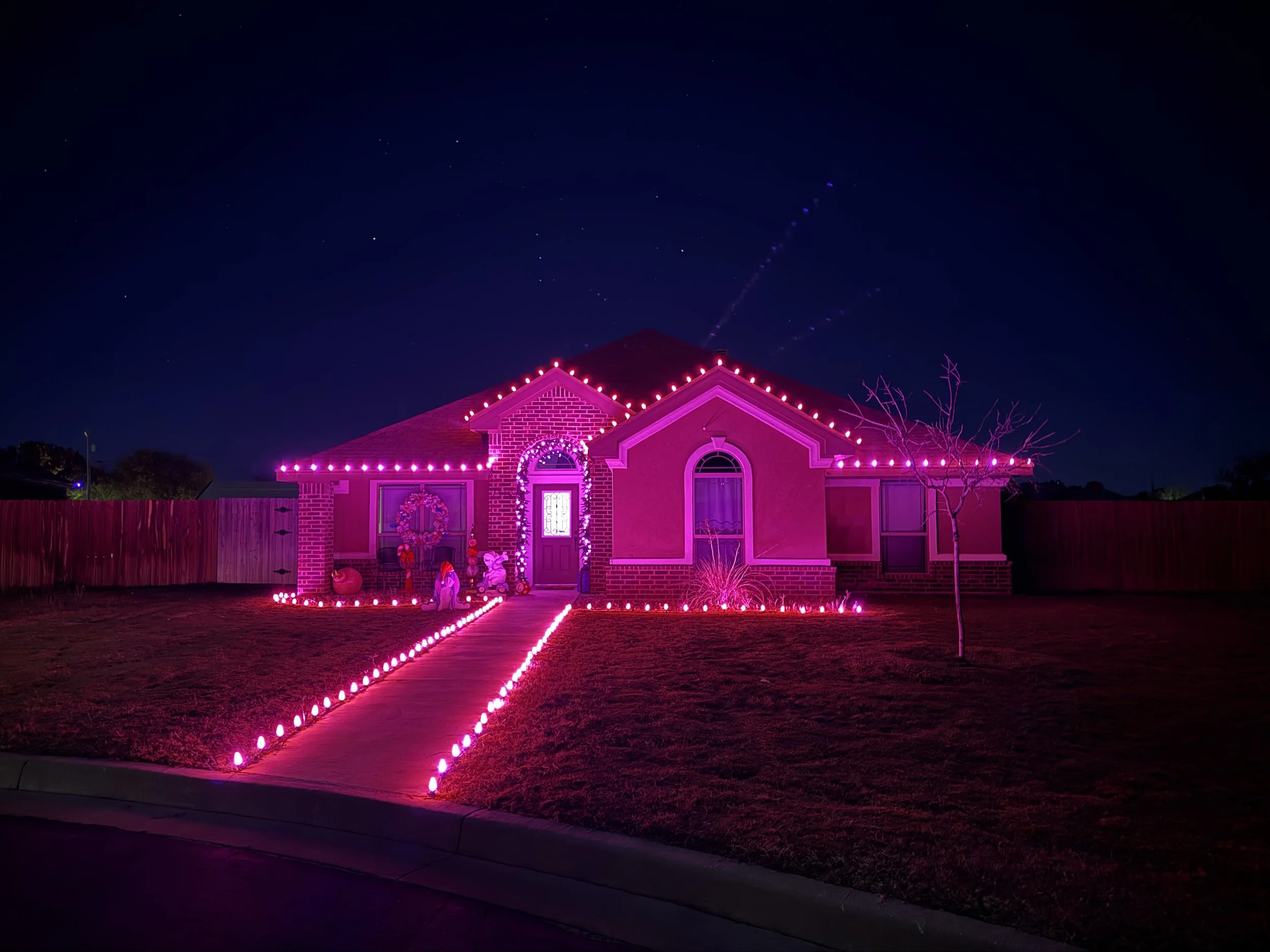 A house decorated with pink Christmas lights outlining the roof, windows, and door, with a pathway lit by small pink lights. The yard features a small tree and holiday decorations in front of the house, against a dark night sky.