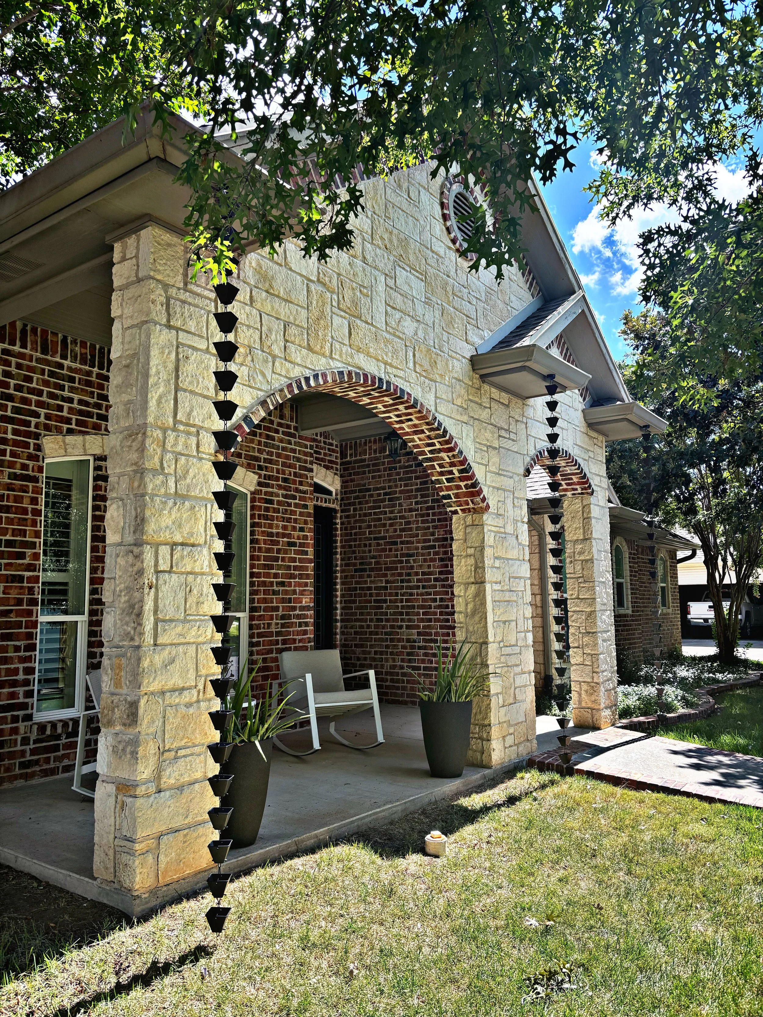 Front porch of a house with stone and brick exterior, with hanging planters, potted plants, and a white bench under a covered porch, surrounded by trees and grass, with a bright blue sky with clouds.