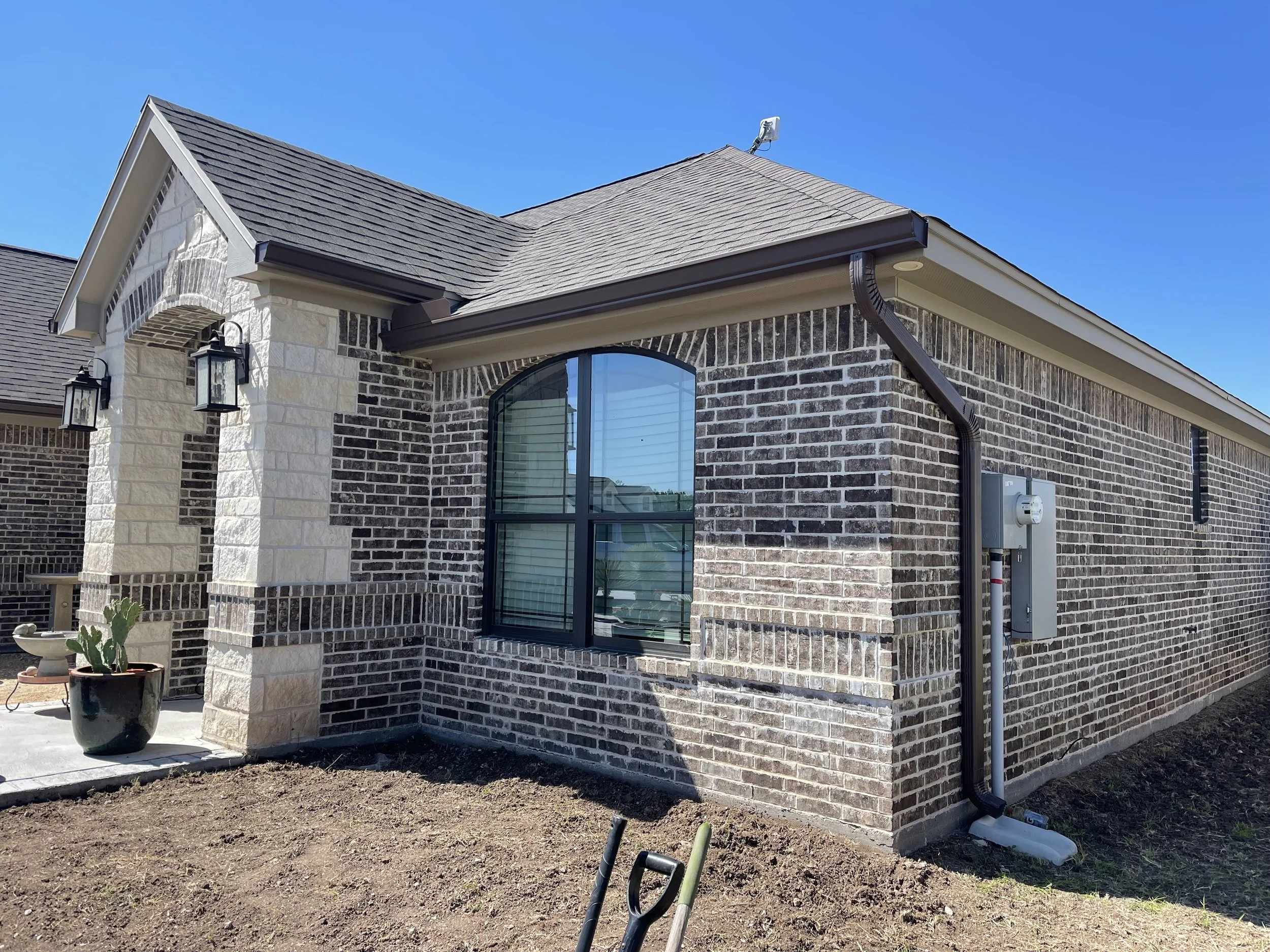 The image shows the exterior corner of a brick house with a grey shingled roof, black gutter, and black window frames. There are two black outdoor wall lanterns near the corner, a potted cactus plant on a concrete patio, and some construction tools a