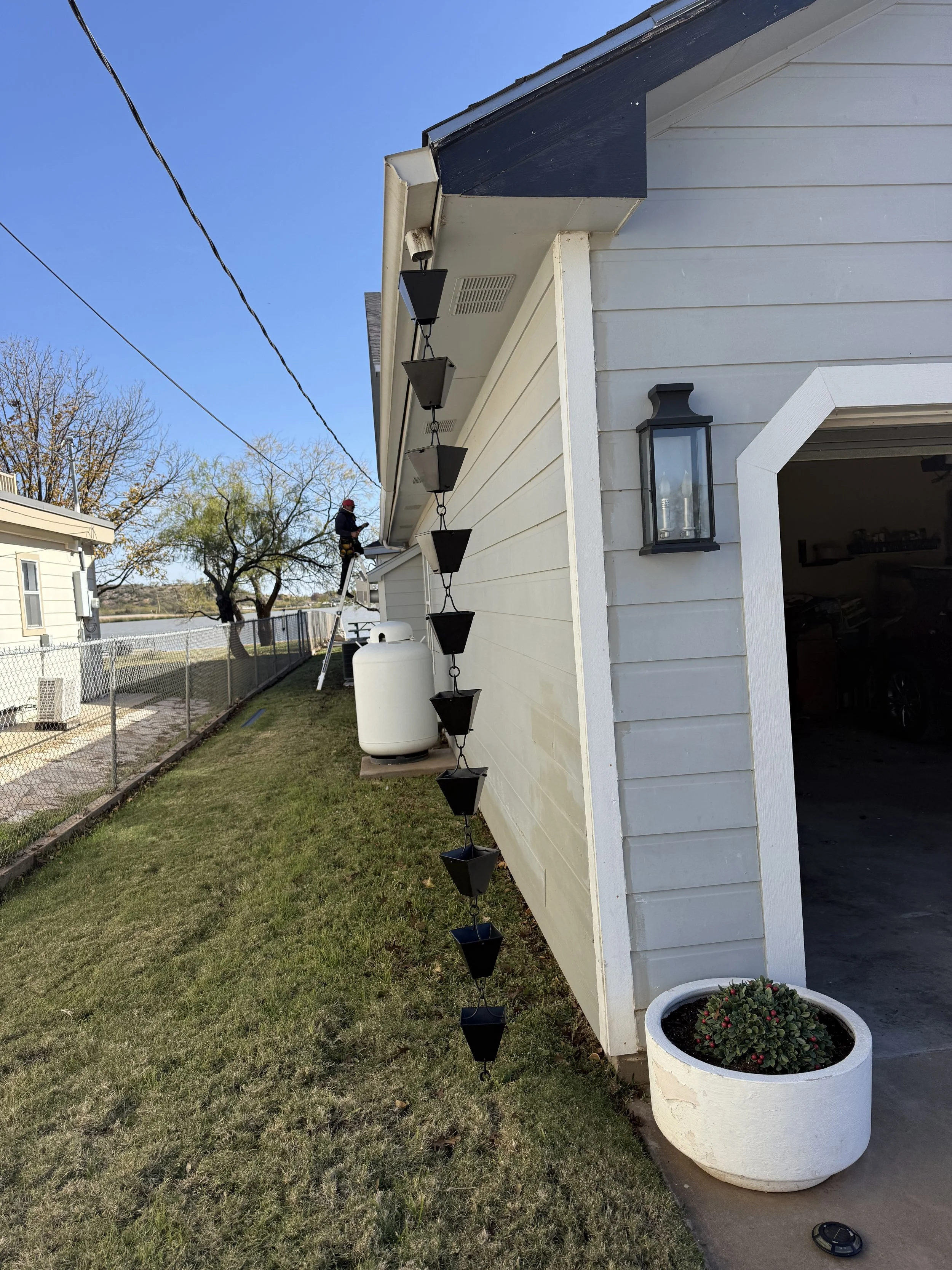 A person standing on a ladder working on the roof of a white house with black trim, under a clear blue sky. The house has outdoor lighting fixtures and a hanging decor with black flower pots. There is a grassy yard with a chain-link fence and a propa