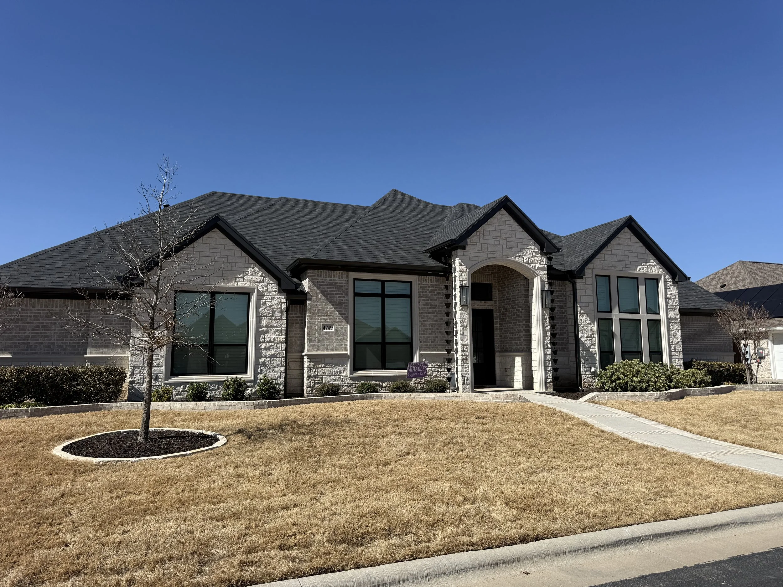 A modern, two-story house with a brick exterior and large windows, situated in a neighborhood with similar homes. The front yard is landscaped with a small tree, bushes, a concrete walkway, and a grass lawn.