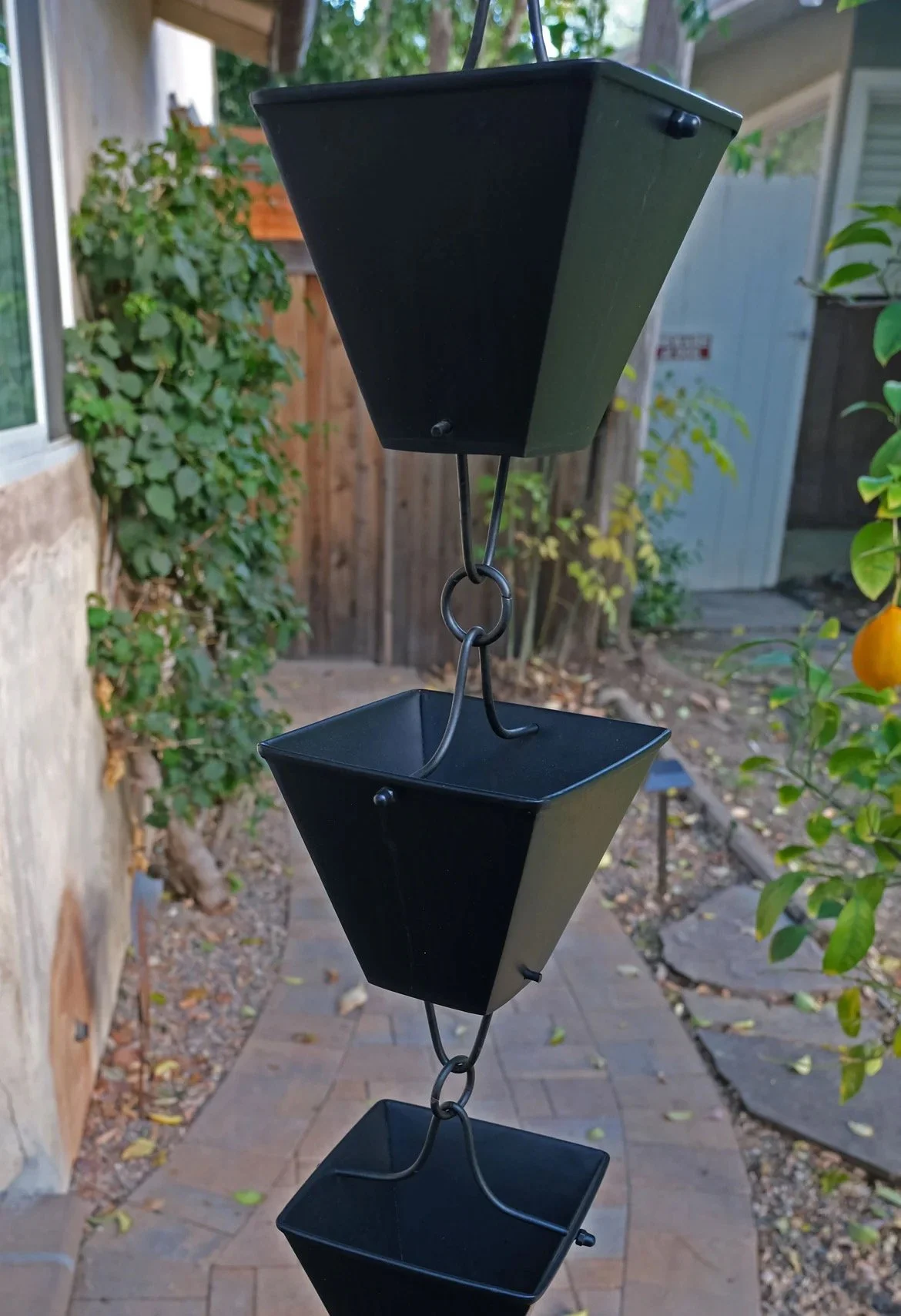A black metal bird feeder with three empty trays hanging in a garden area with plants, a brick pathway, and a fence in the background.