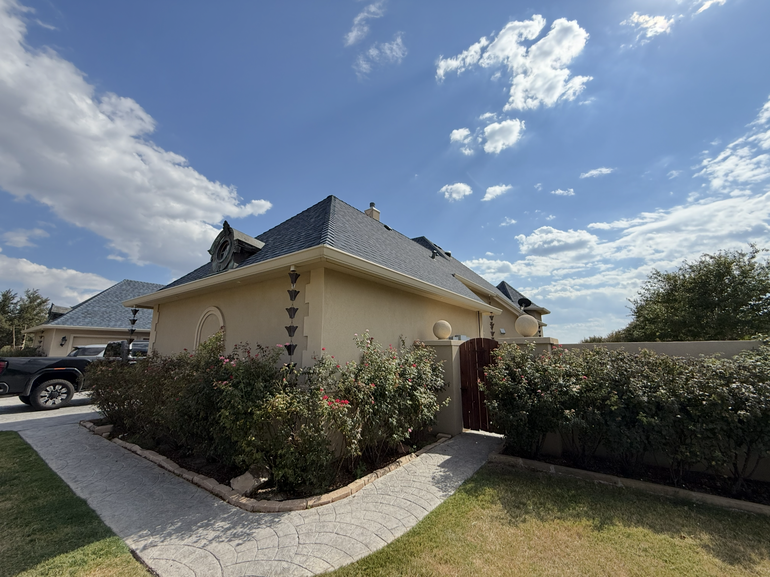 A house with a gray shingle roof under a partly cloudy blue sky, front yard with flowers and bushes, parked cars outside.