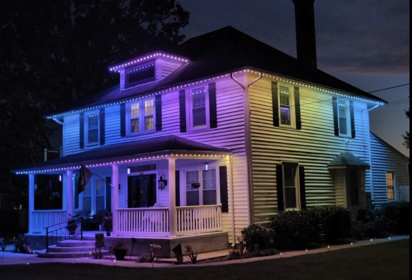 A multi-story house decorated with colorful string lights at night. The house has a porch, several windows with shutters, and a dark roof. The lights create a rainbow effect around the house's edges.