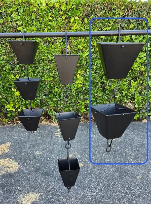Six black hanging planters in a row, suspended from a metal rod, with a background of green bushes and a gray asphalt ground.