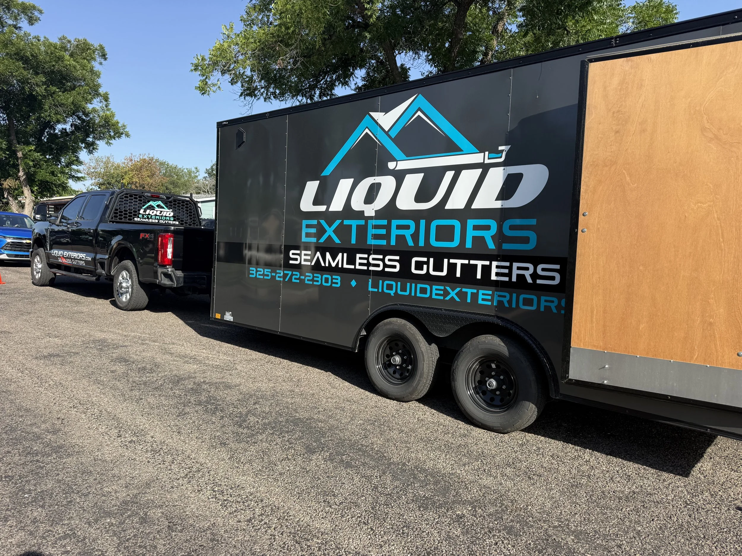 A black truck and trailer parked outdoors. The trailer has the logo and text for Liquid Exteriors Seamless Gutters. Trees and a clear blue sky are in the background.