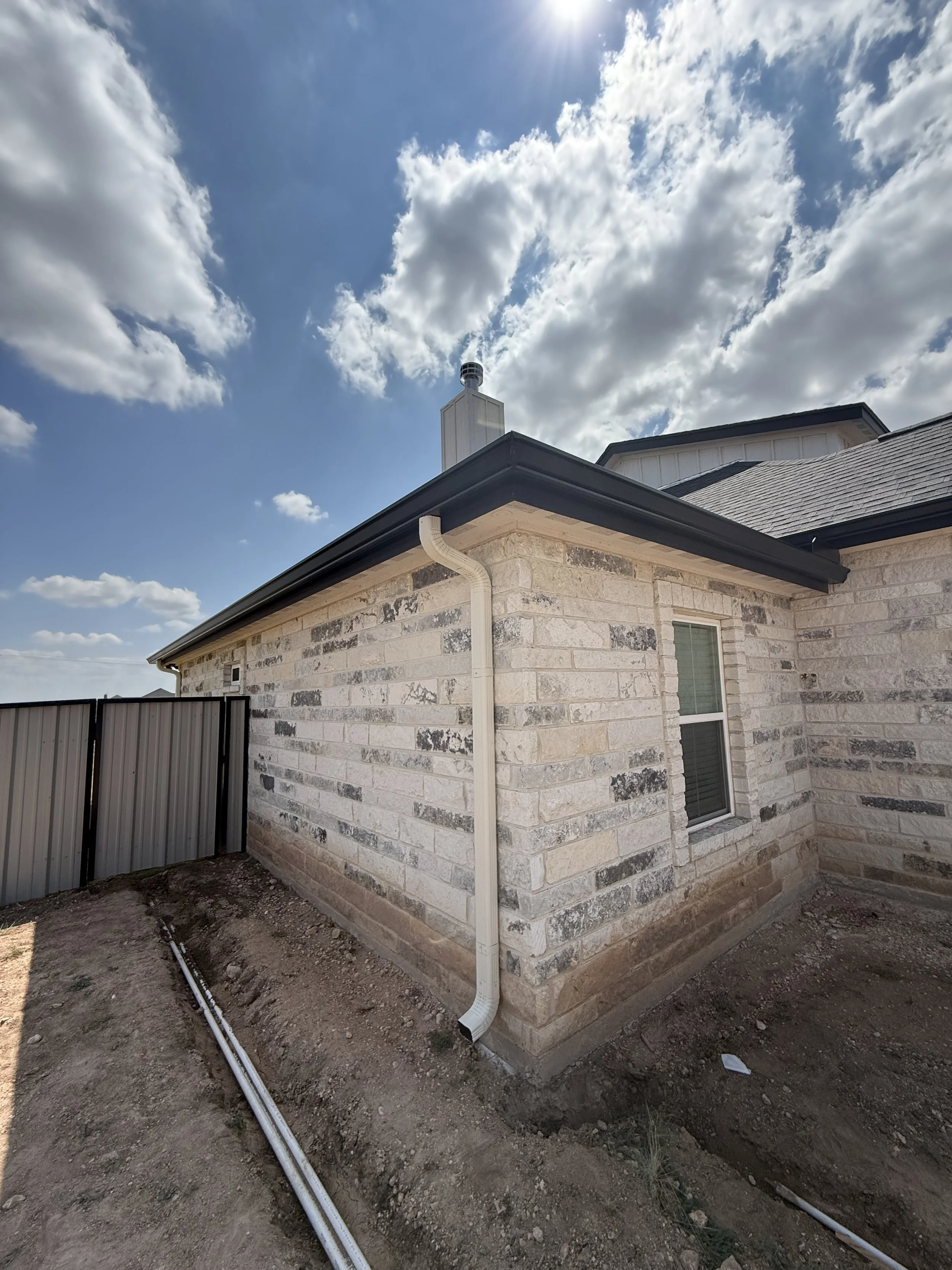 The exterior of a house with a stone brick wall, a black roof, and a white downspout, under a partly cloudy sky with sunlight.