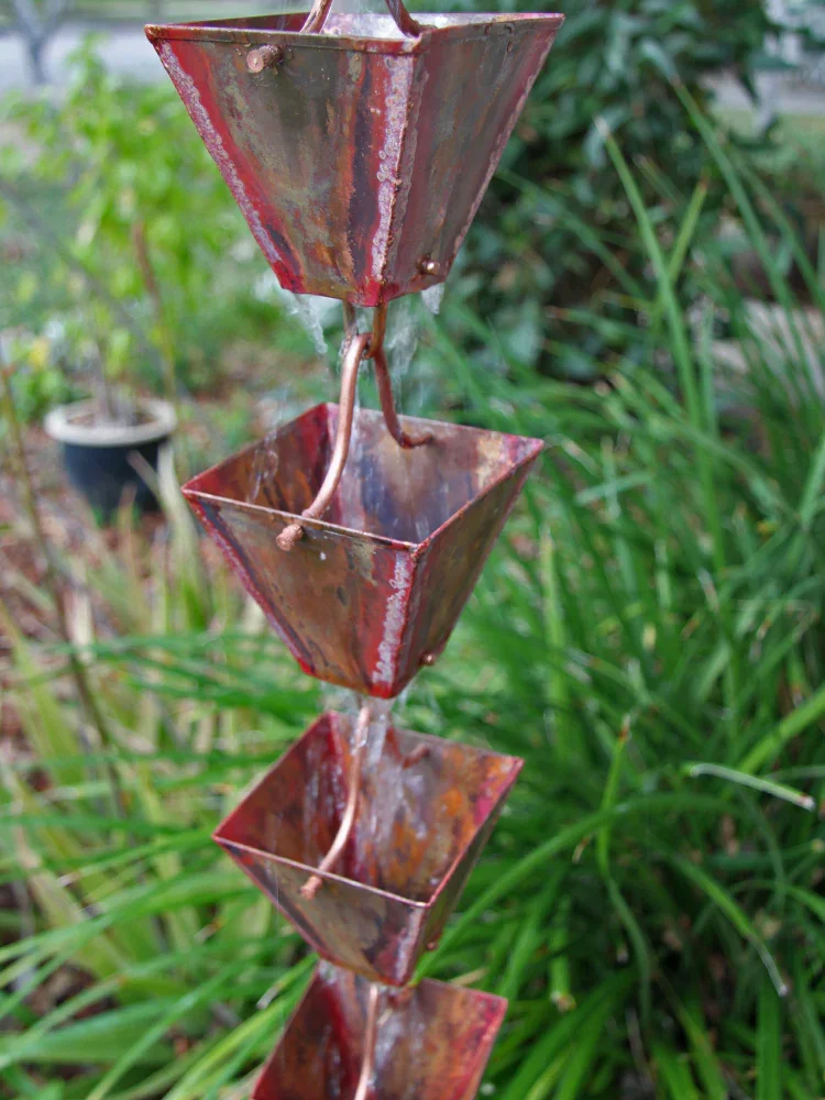 Close-up of a vertical rusted metal water fountain with four small square basins, with water flowing from the top basin to the others, set against green plants in the background.
