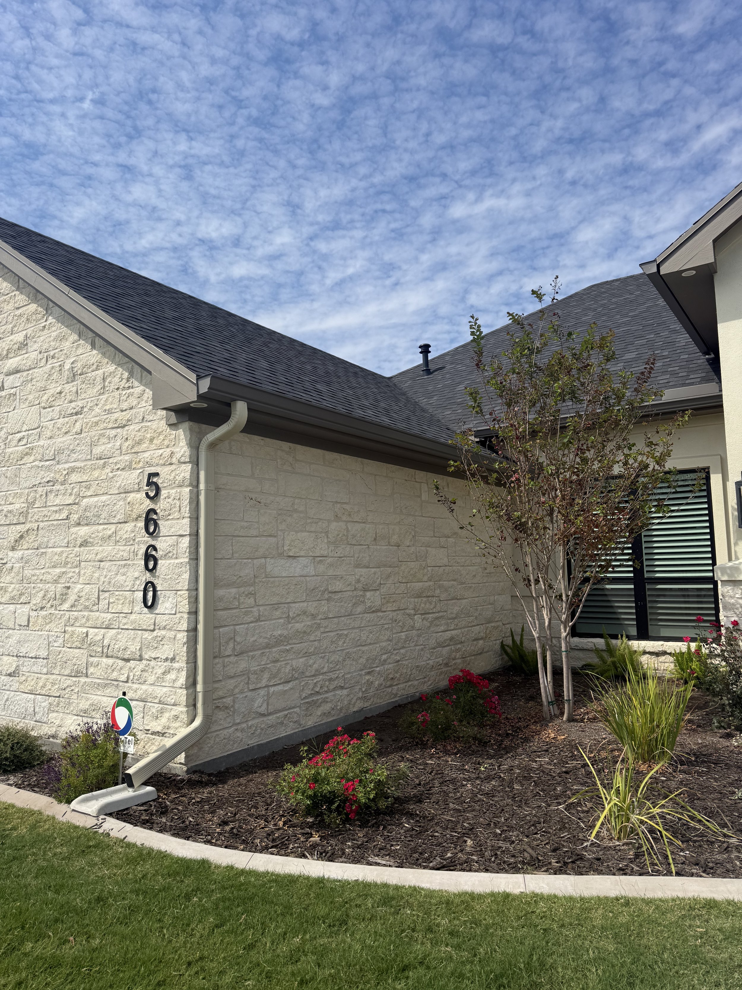 Exterior view of a house with a textured white brick wall, black roof, and a landscaped garden with flowers and trees