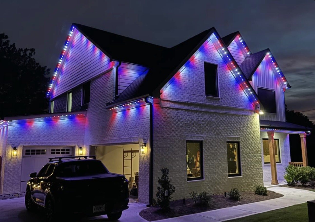 A two-story house lit with colorful Christmas lights at night. The house has a garage with a black SUV parked outside, and the front porch has wooden columns and shrubs nearby.