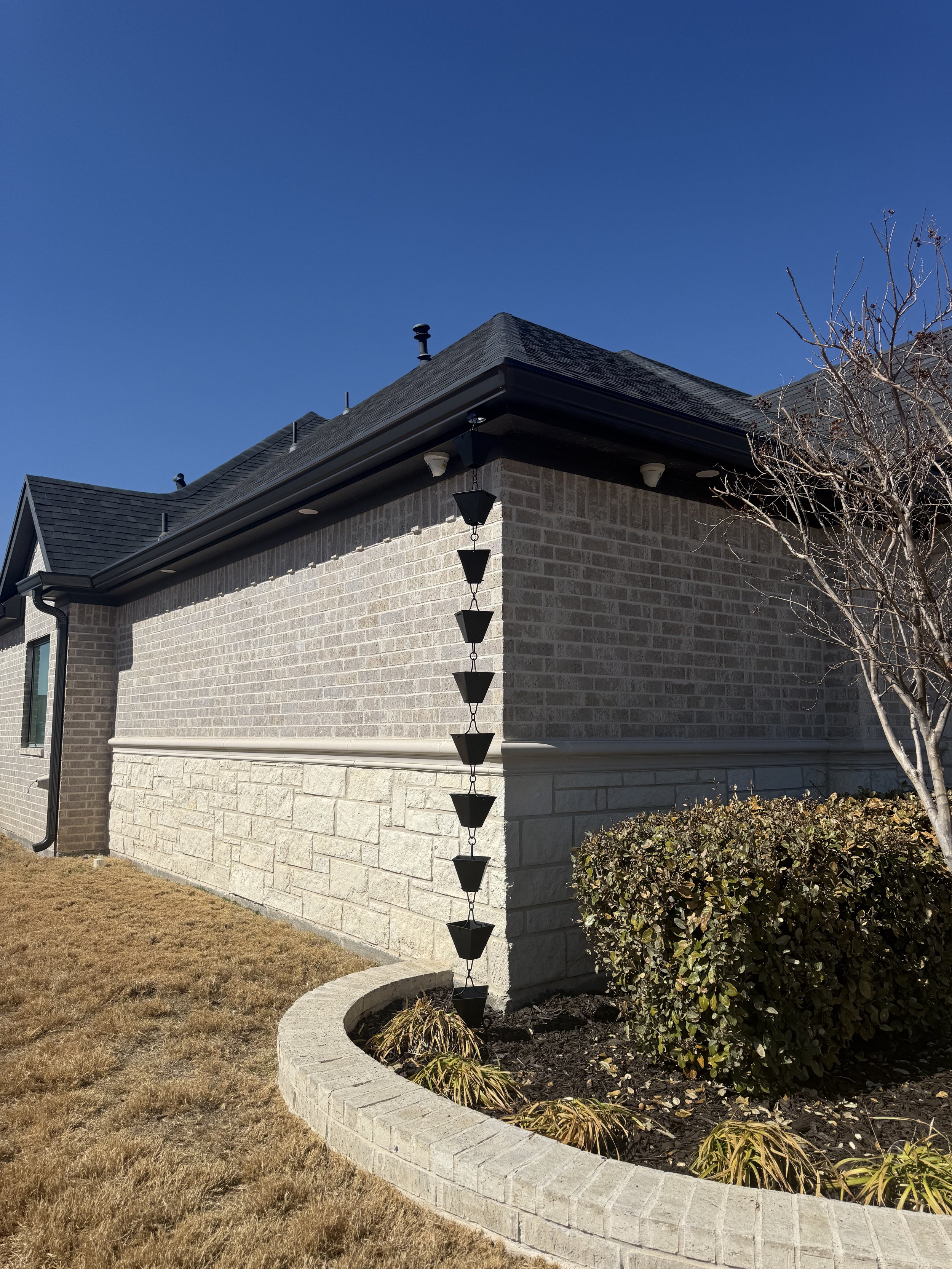 Corner of a house with brick and stone exterior, black roof shingles, black gutter system, decorative black lantern, leafless tree, and a shrub in a curved brick flower bed, under a clear blue sky.