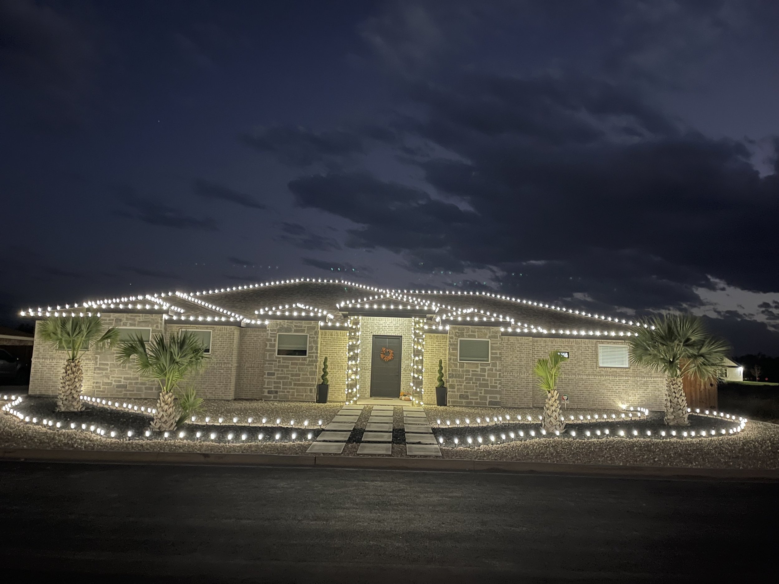 A house decorated with white string lights outlining the roof, front porch, and walkway, with palm trees in the front yard and a cloudy night sky overhead.