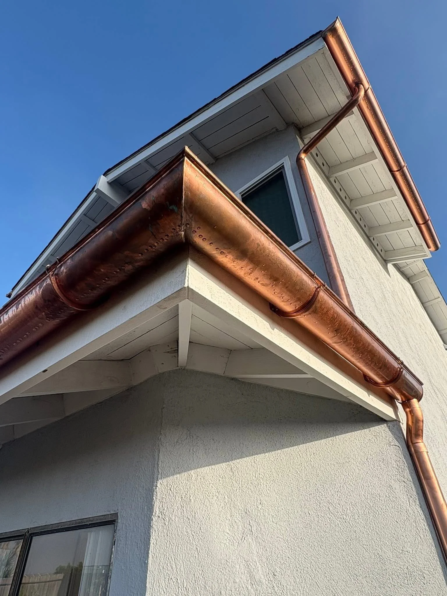 Close-up of a house corner with white stucco walls, a small window, and a copper gutter system against a clear blue sky.