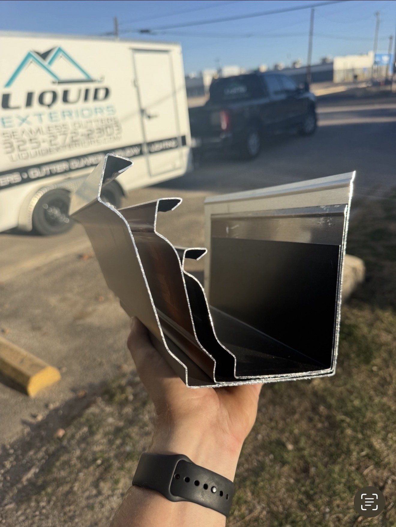 Person holding a piece of aluminum framing in front of a parking lot with vehicles, including a truck with liquid exterior signage.
