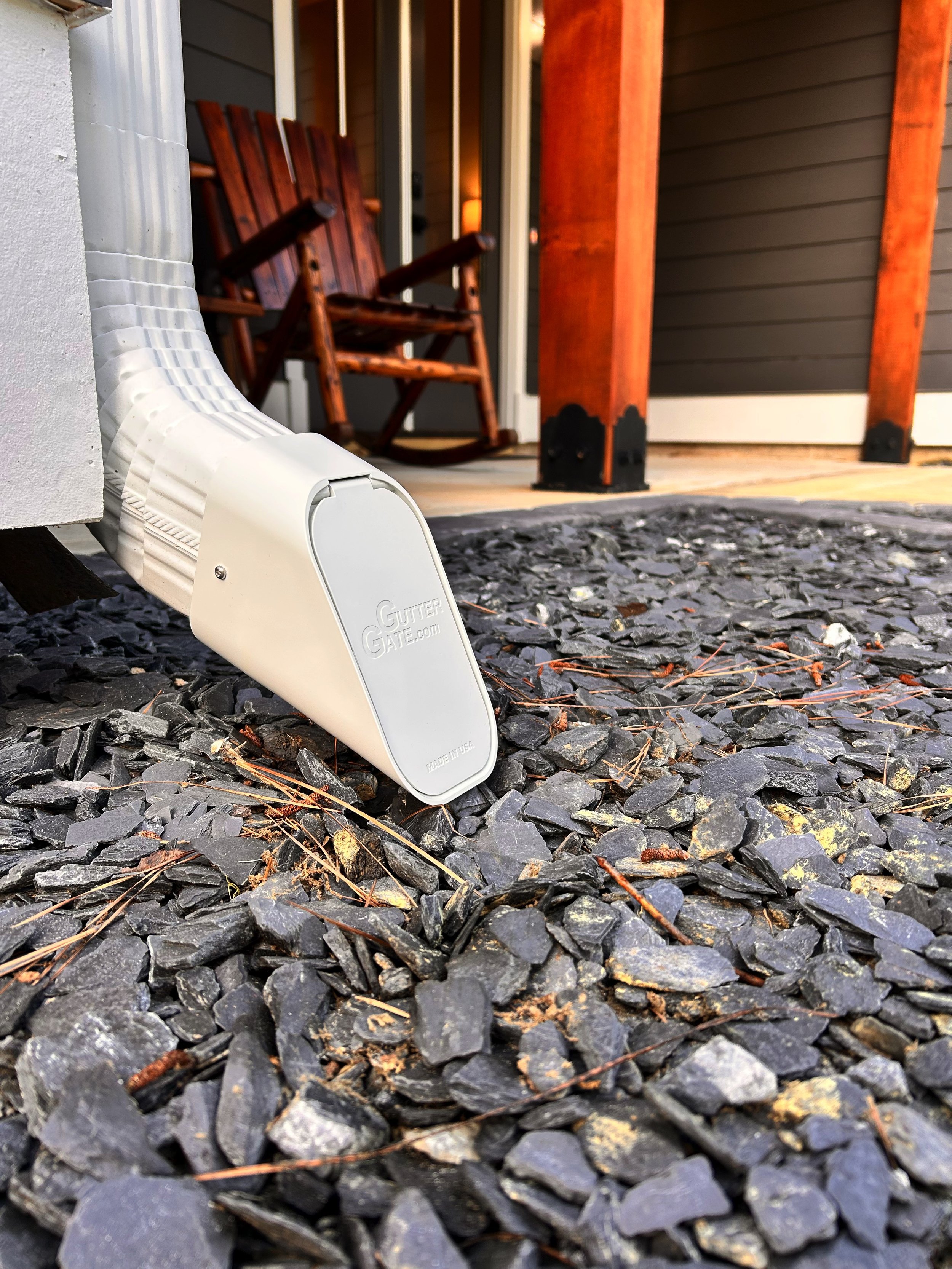 Close-up of a white gutter downspout extending onto a rocky surface in front of a porch with a wooden rocking chair and porch furniture.