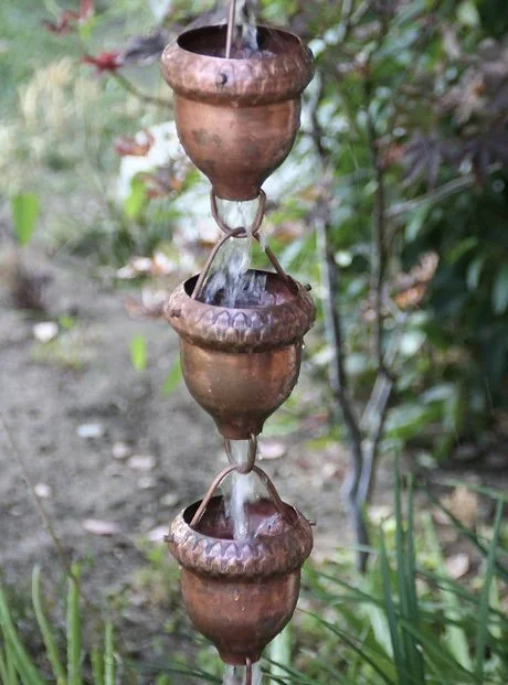 A vertical string of three copper water containers with water flowing from the top container to the middle, then to the bottom, surrounded by greenery and garden plants.
