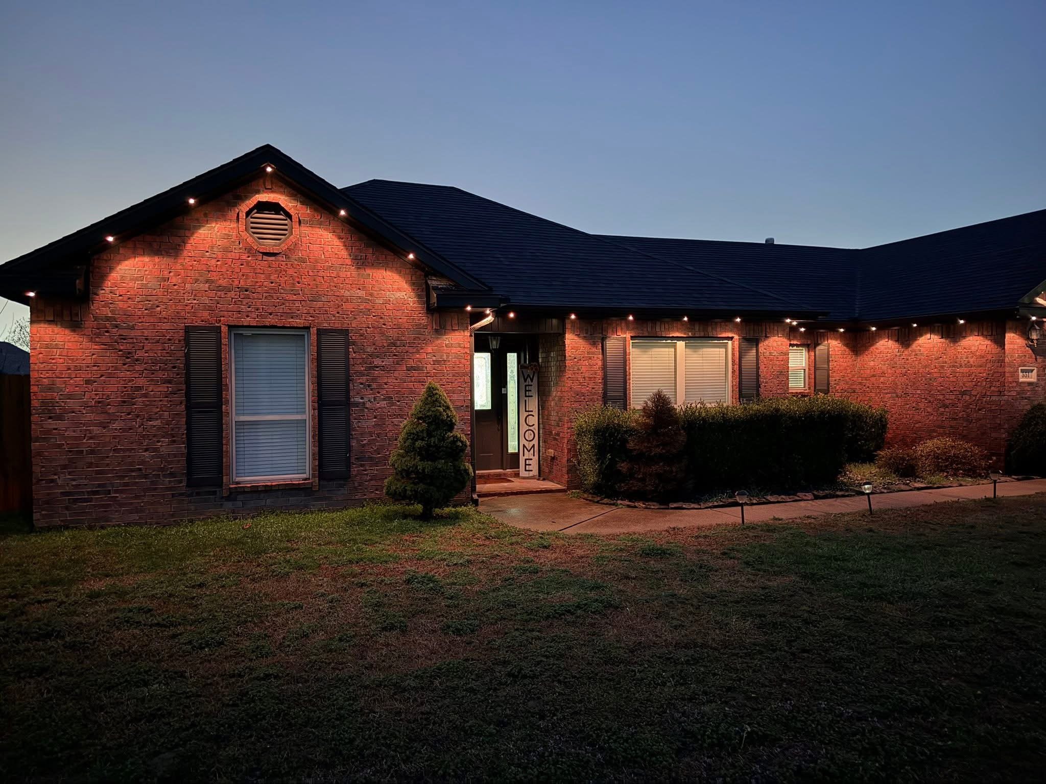 A brick house with a dark roof illuminated by exterior string lights at dusk or early evening.