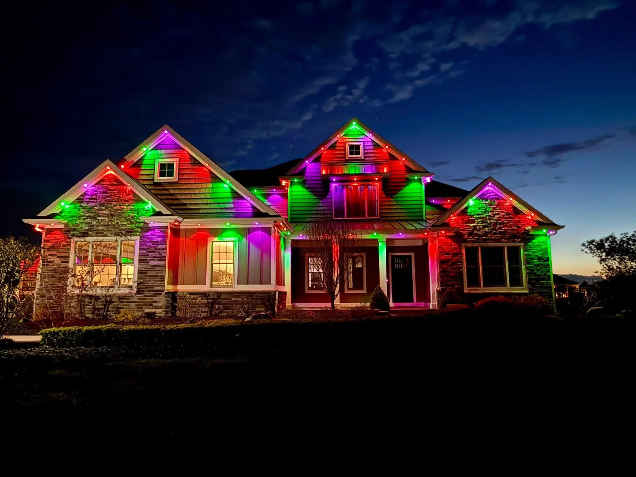 A house decorated with colorful Christmas lights at night, with glowing red, green, purple, and pink lights outlining the roof and windows, against a dark sky with some clouds.