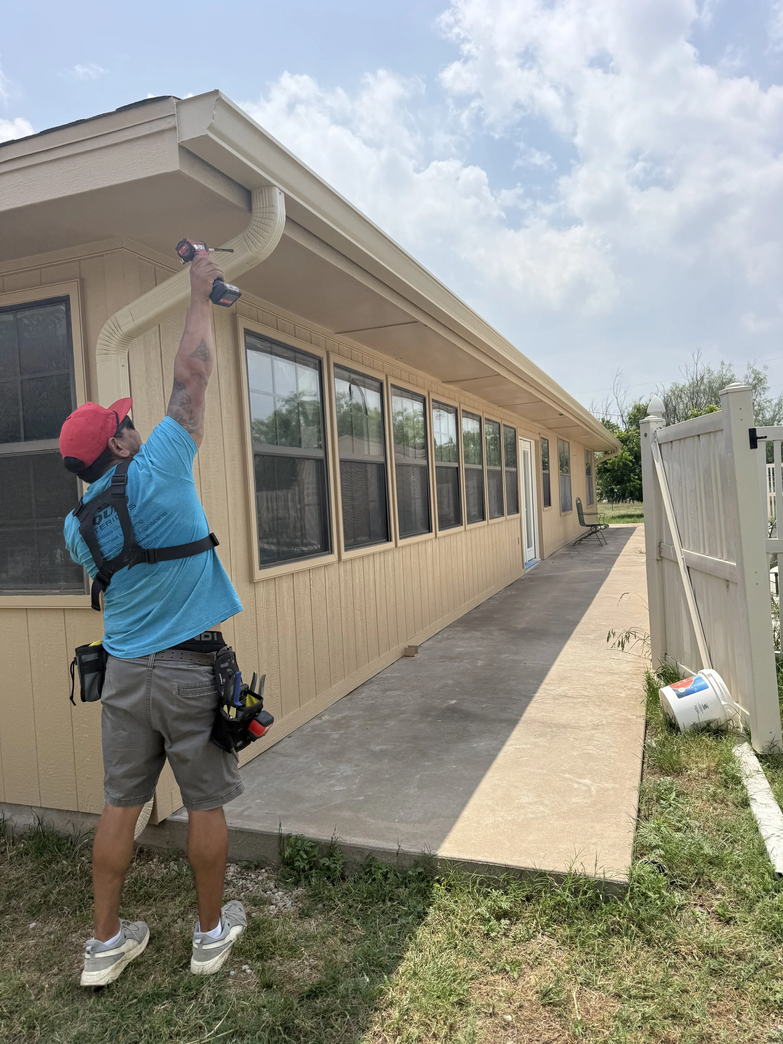 A man wearing a red cap, blue t-shirt, grey shorts, and sneakers is working outside on a beige house's gutter system, using a cordless drill. The house has large windows, and a white fence is visible on the right. The sky is partly cloudy, and there is some grass and weeds near the concrete patio.