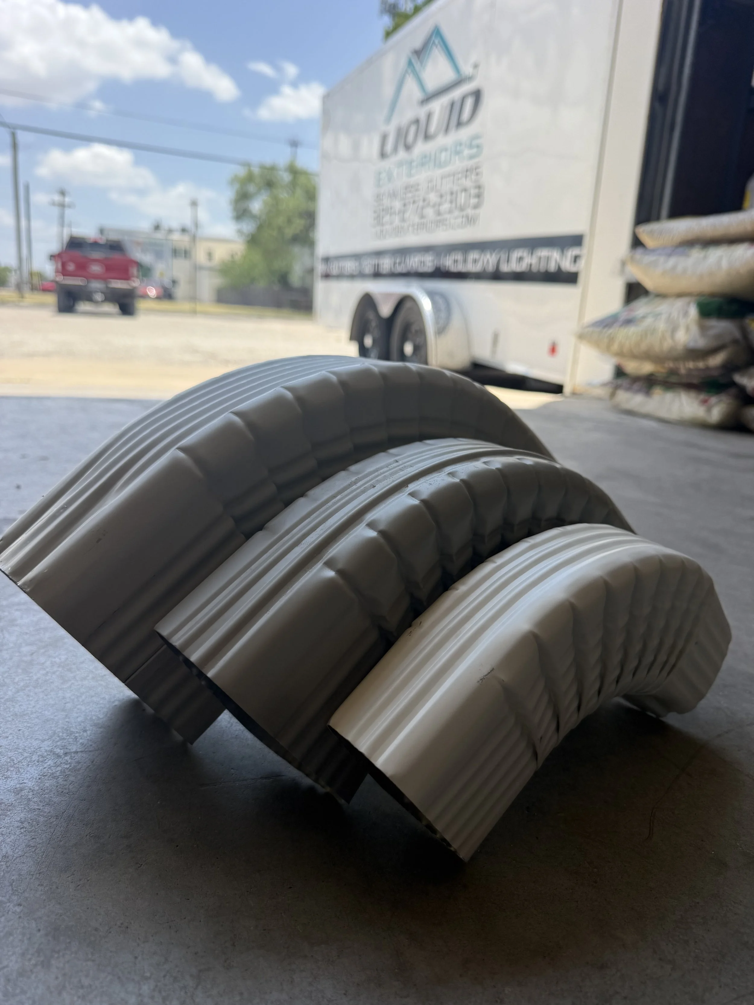 A close-up view of several rolled-up black and gray flexible hoses or semi-rigid pipes lying on a concrete floor. In the background, there is a white trailer with the words "Liquid Interiors" and related services printed on it, along with a red picku
