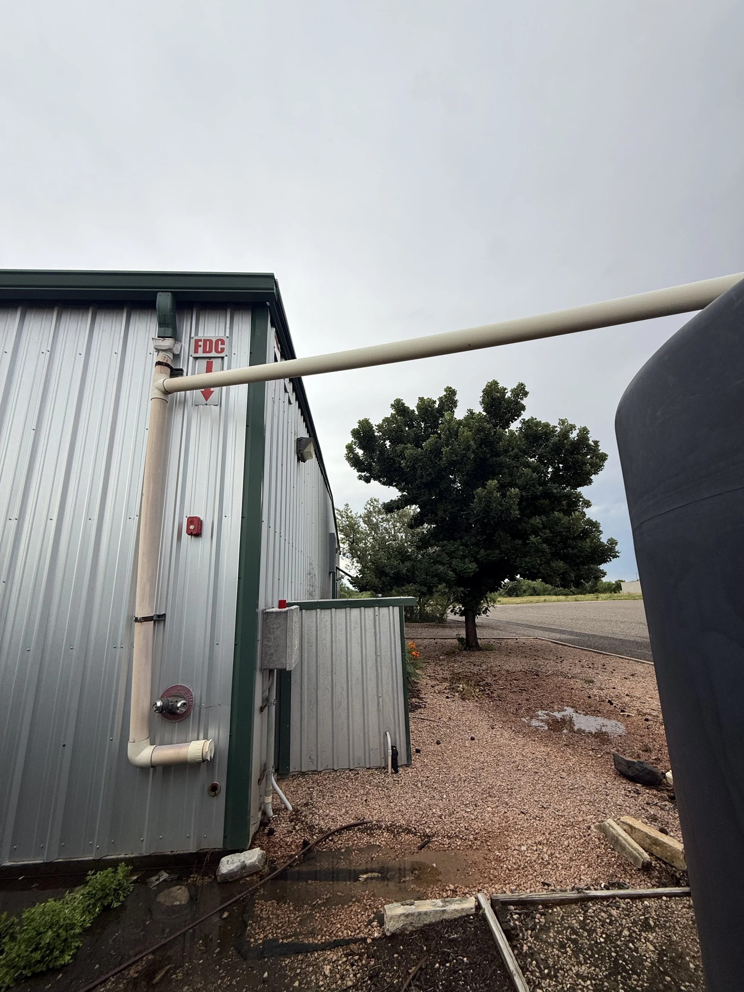 Outdoor view of a metal building with piping and a tree in the background, overcast sky.